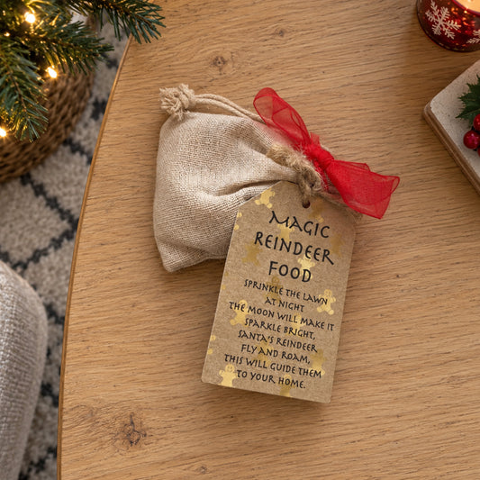 A small beige burlap drawstring pouch tied with a red sheer ribbon and a kraft paper tag that reads 'Magic Reindeer Food' with a humorous holiday instruction, placed on a wooden table.