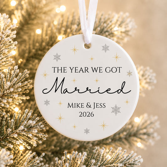 A round white Christmas ornament with a white hanging ribbon, printed with the text 'THE YEAR WE GOT Married MIKE & JESS 2026' in black lettering, decorated with small gold and silver star and snowflake motifs. The ornament is shown hanging in front of a softly blurred festive Christmas tree with warm lights.