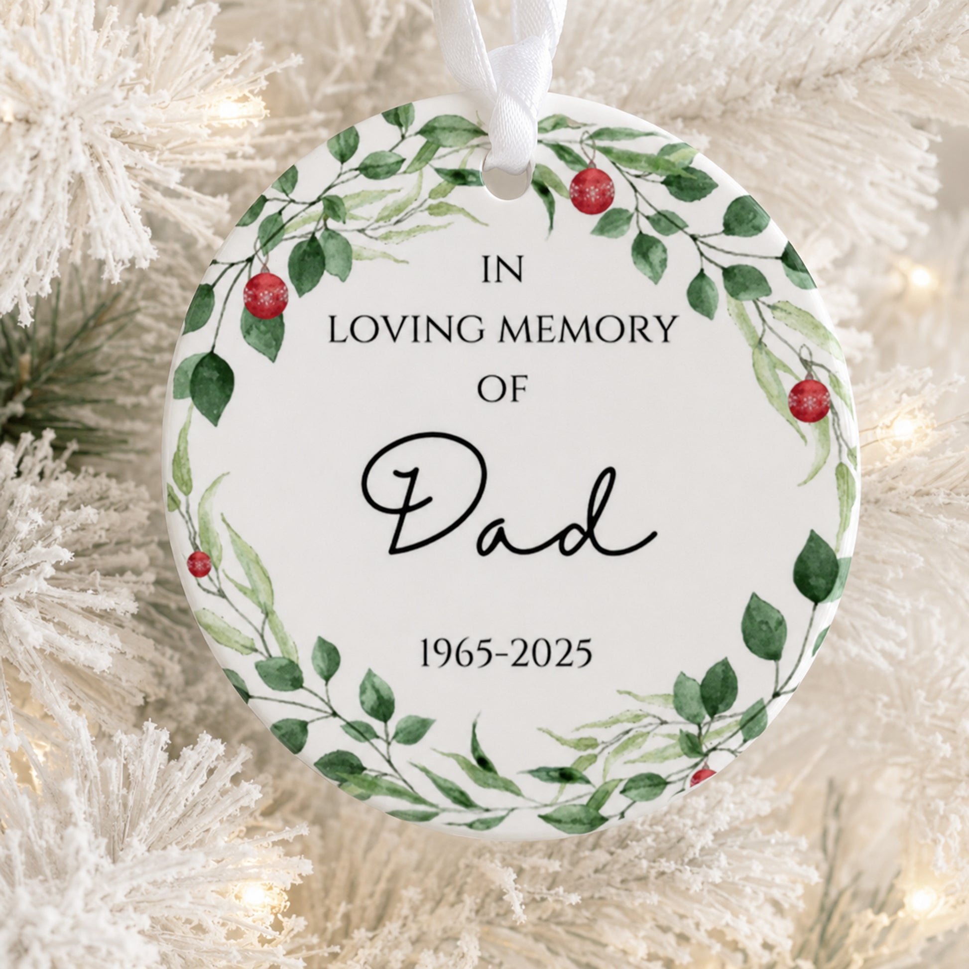A round white Christmas ornament with a watercolor green leaf wreath and red berries, hanging on a white frosted Christmas tree. The ornament features the printed text 'IN LOVING MEMORY OF Dad 1965-2025' in black lettering.