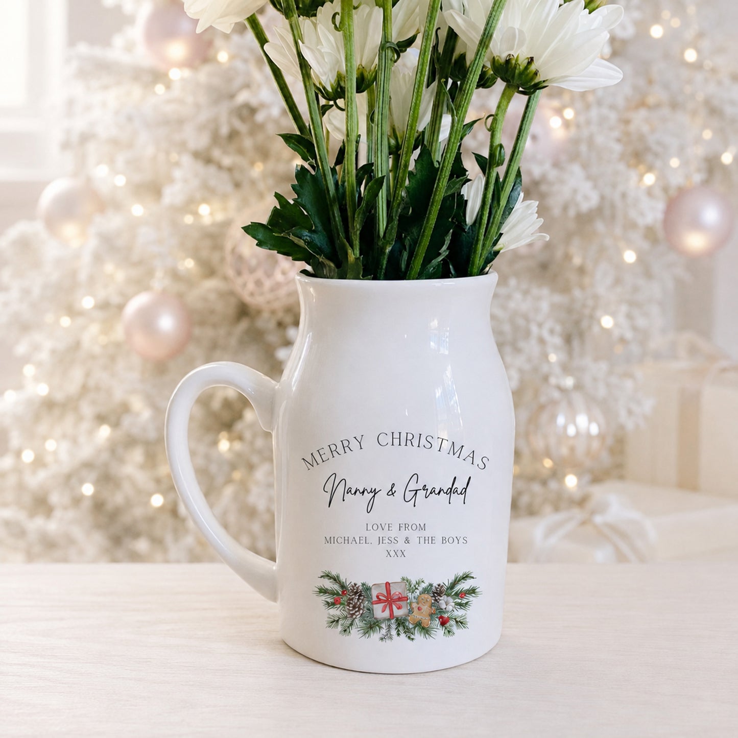 A white ceramic flower jug designed as a pitcher, filled with white daisies, set against a festive blurred Christmas tree background. The mug features the text 'MERRY CHRISTMAS Man & Grand' with a personalized message below and a small holiday wreath illustration at the bottom.