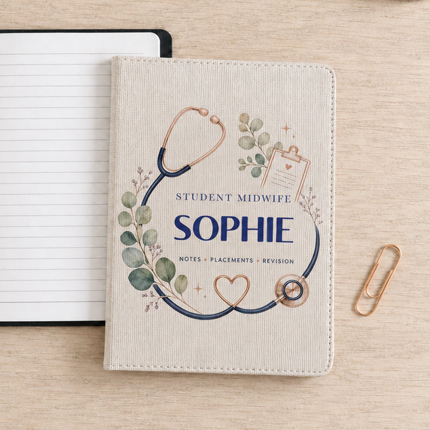 A personalized cream-colored leather notebook cover with a stethoscope and eucalyptus design, featuring the text 'STUDENT MIDWIFE SOPHIE' and 'NOTES - PLACEMENTS - REVISION'. It is displayed on a light wood desk next to an open lined notebook, a pen, and a gold paperclip.