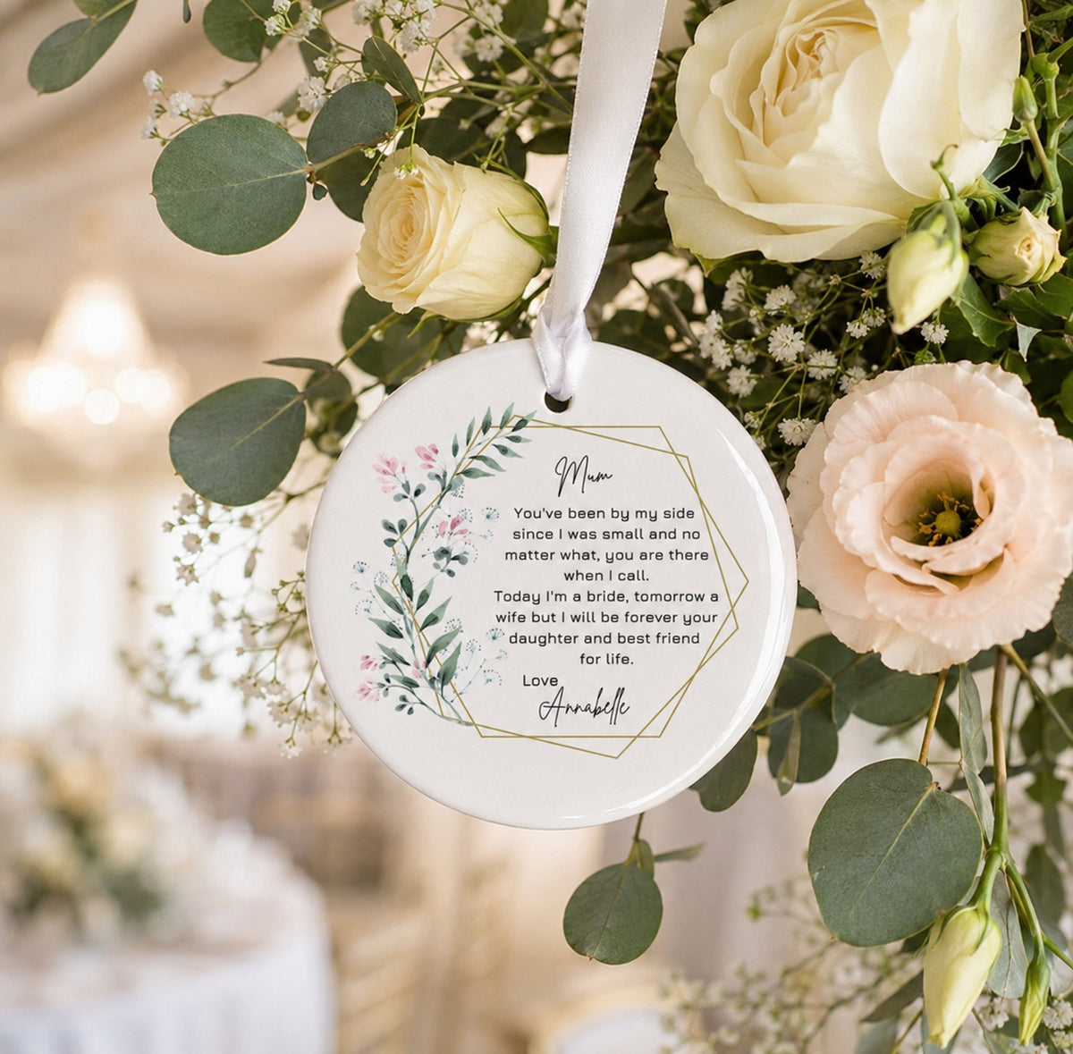 A round white ceramic wedding keepsake ornament with a floral wreath design and a printed sentimental message for 'Mum,' from the bride, hanging from a white ribbon amidst cream roses, pale peach flowers, and green eucalyptus leaves.
