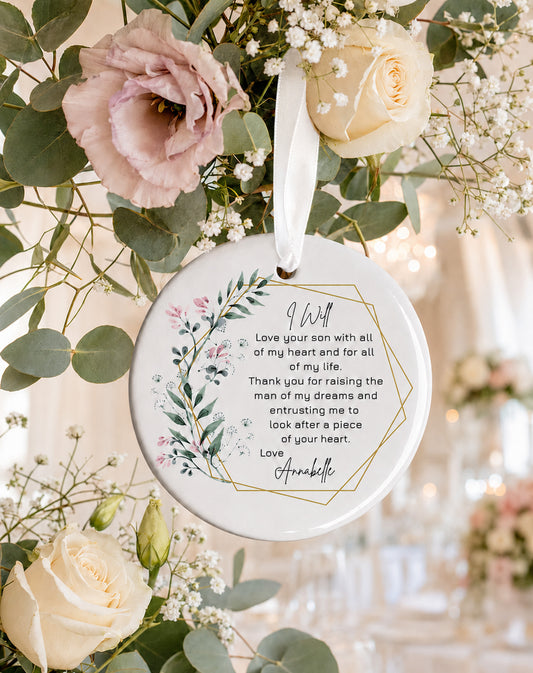 A round white ceramic ornament with a printed sentimental message for the Mother of the Groom,  from the Bride hanging from a white ribbon amidst floral arrangements with pink roses and green eucalyptus