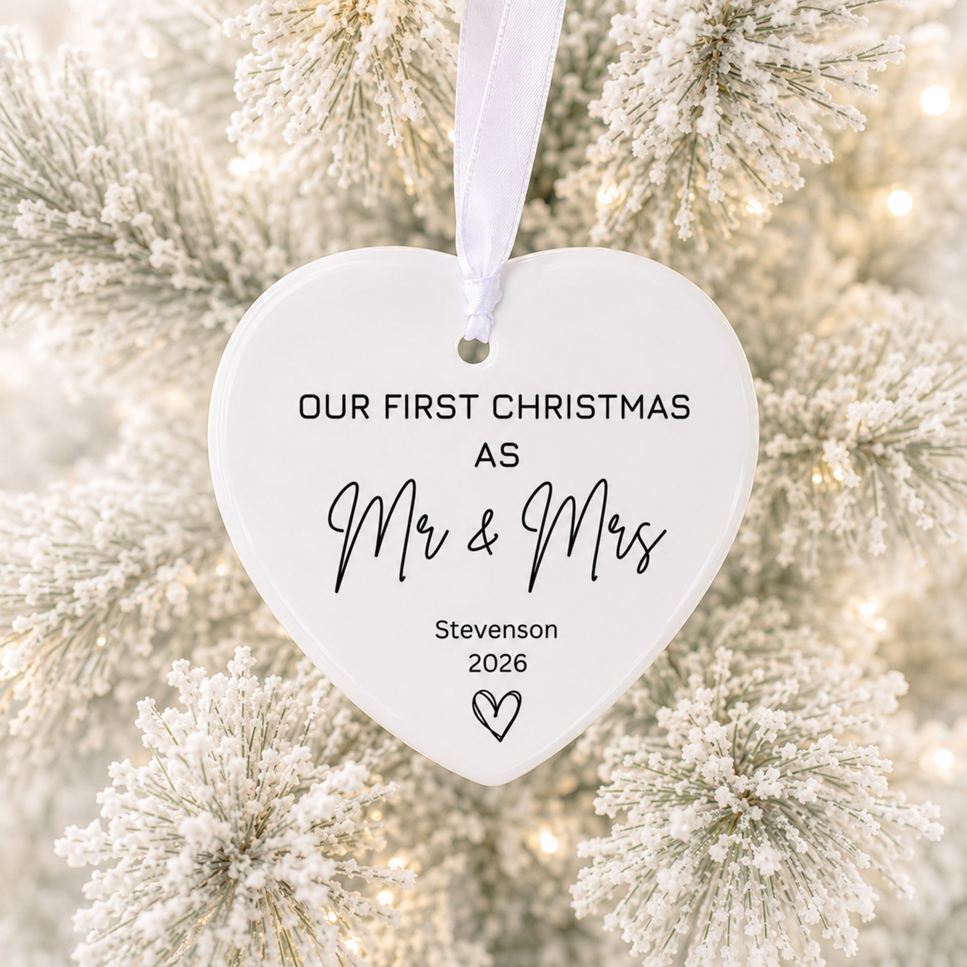 A white heart-shaped ceramic Christmas ornament hanging from a white ribbon, printed with the text 'OUR FIRST CHRISTMAS AS Mr & Mrs Stevenson 2026' and a small heart, set against a blurred background of frosted pine branches and warm string lights.