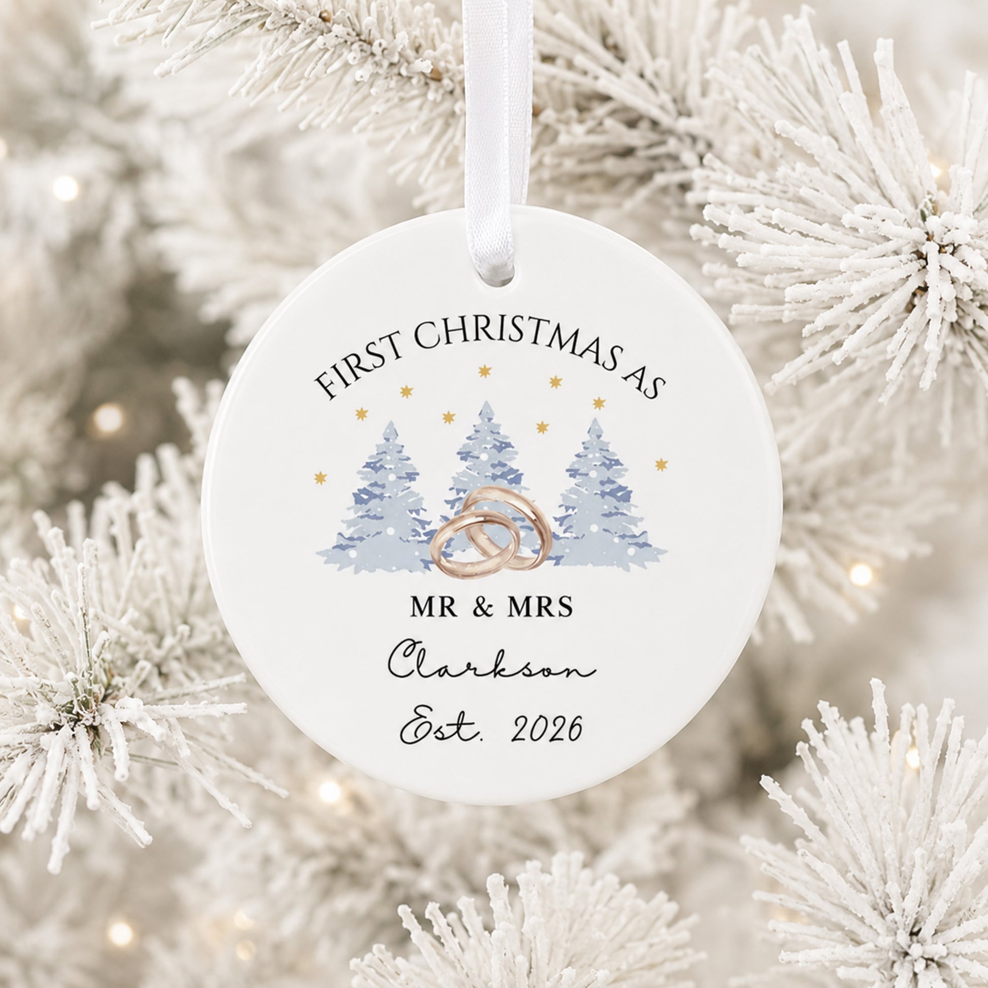A round white Christmas ornament hanging from a white ribbon, printed with the text 'FIRST CHRISTMAS AS MR & MRS Clarkson Est. 2026' and an illustration of three blue pine trees with interlocking rings.