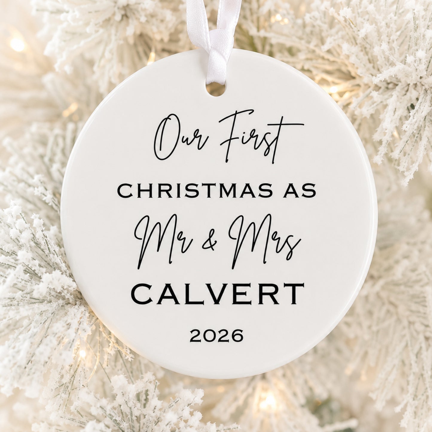 A round white ceramic Christmas ornament hanging from a white ribbon, printed with the personalized text 'Our First Christmas As Mr & Mrs CALVERT 2026' in black lettering, set against a blurred background of a white-frosted Christmas tree with warm lights.