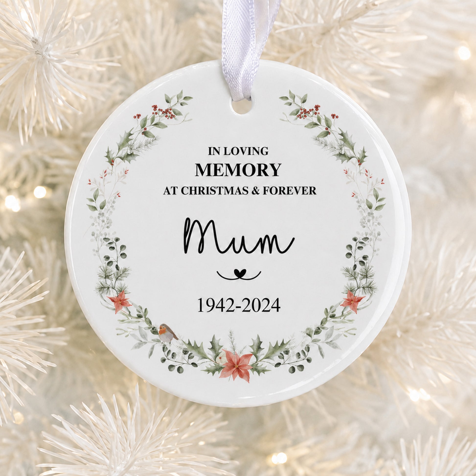 A round white ceramic Christmas memory ornament hanging from a white ribbon, printed with the text 'IN LOVING MEMORY AT CHRISTMAS & FOREVER Mum 1942-2024' and decorated with a floral wreath border, shown against a blurred white Christmas tree background.