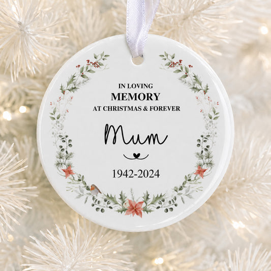 A round white ceramic Christmas memory ornament hanging from a white ribbon, printed with the text 'IN LOVING MEMORY AT CHRISTMAS & FOREVER Mum 1942-2024' and decorated with a floral wreath border, shown against a blurred white Christmas tree background.