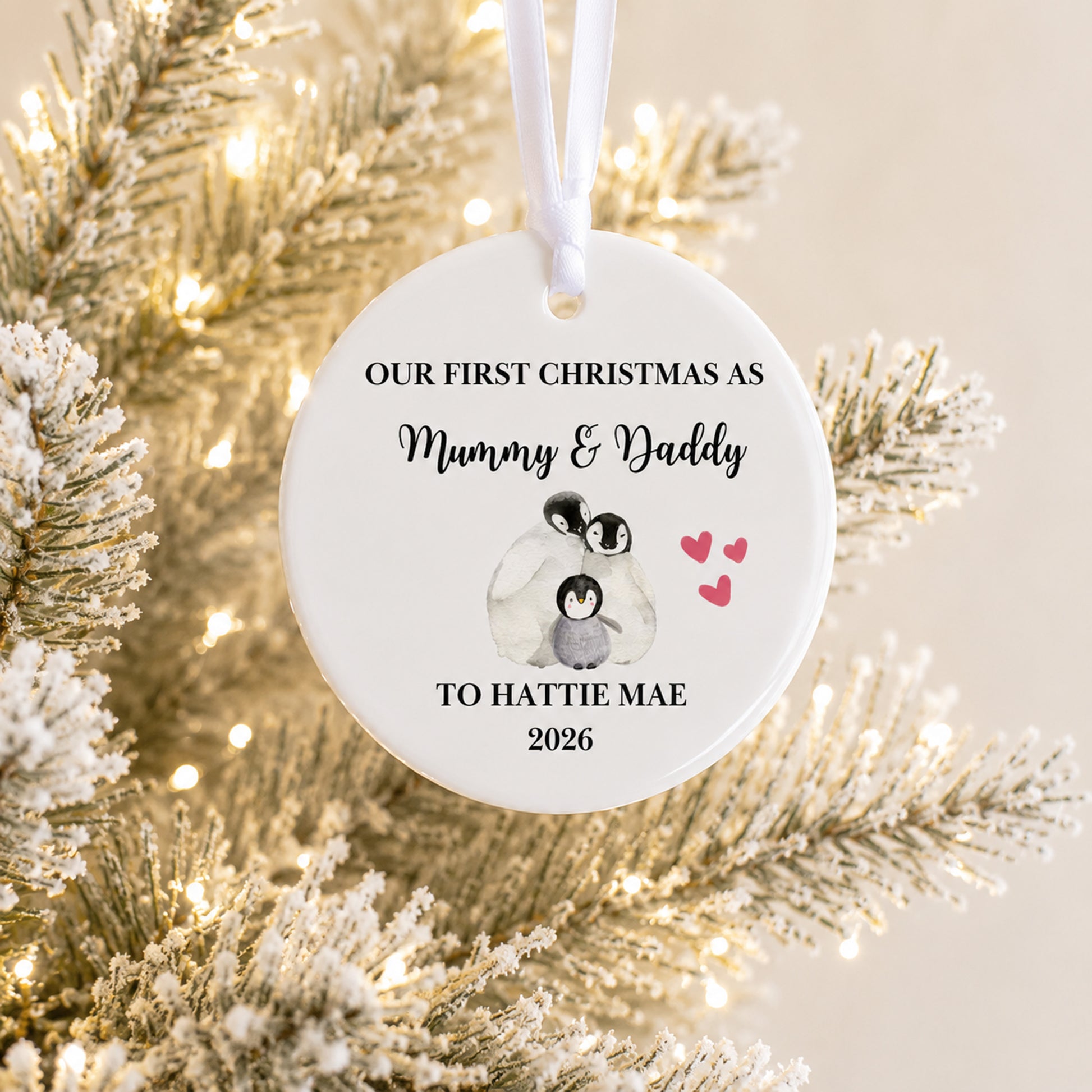 A round white Christmas ornament hanging from a white ribbon, printed with the text 'OUR FIRST CHRISTMAS AS Mummy & Daddy TO HATTIE MAE 2026' and a small illustration of two penguins with pink hearts. The ornament is photographed in front of frosted evergreen branches with warm string lights.