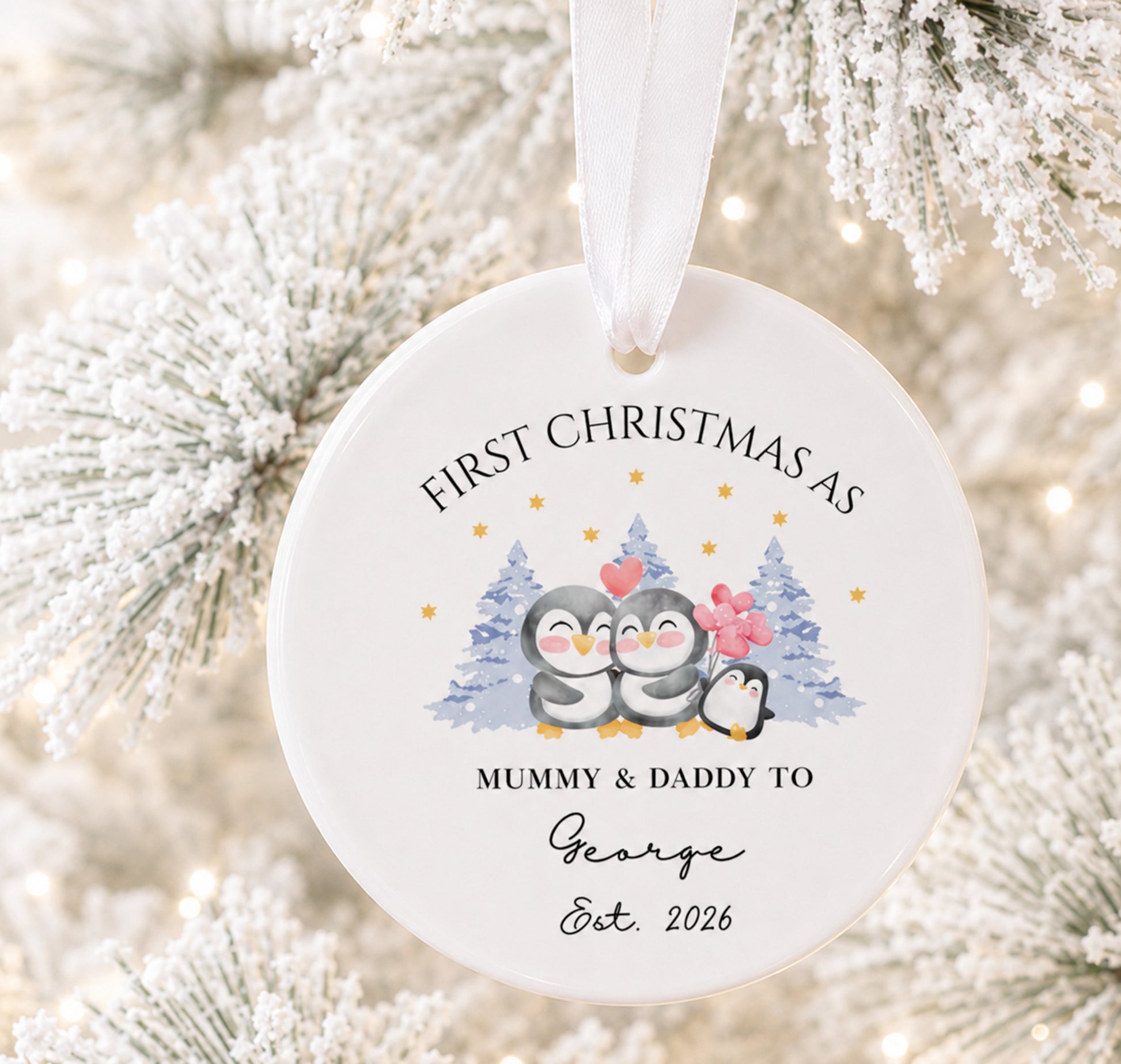 A round white ceramic Christmas ornament hanging from a white ribbon, printed with a penguin illustration and the text 'FIRST CHRISTMAS AS MUMMY & DADDY TO George Est. 2026', set against a blurred flocked Christmas tree background.