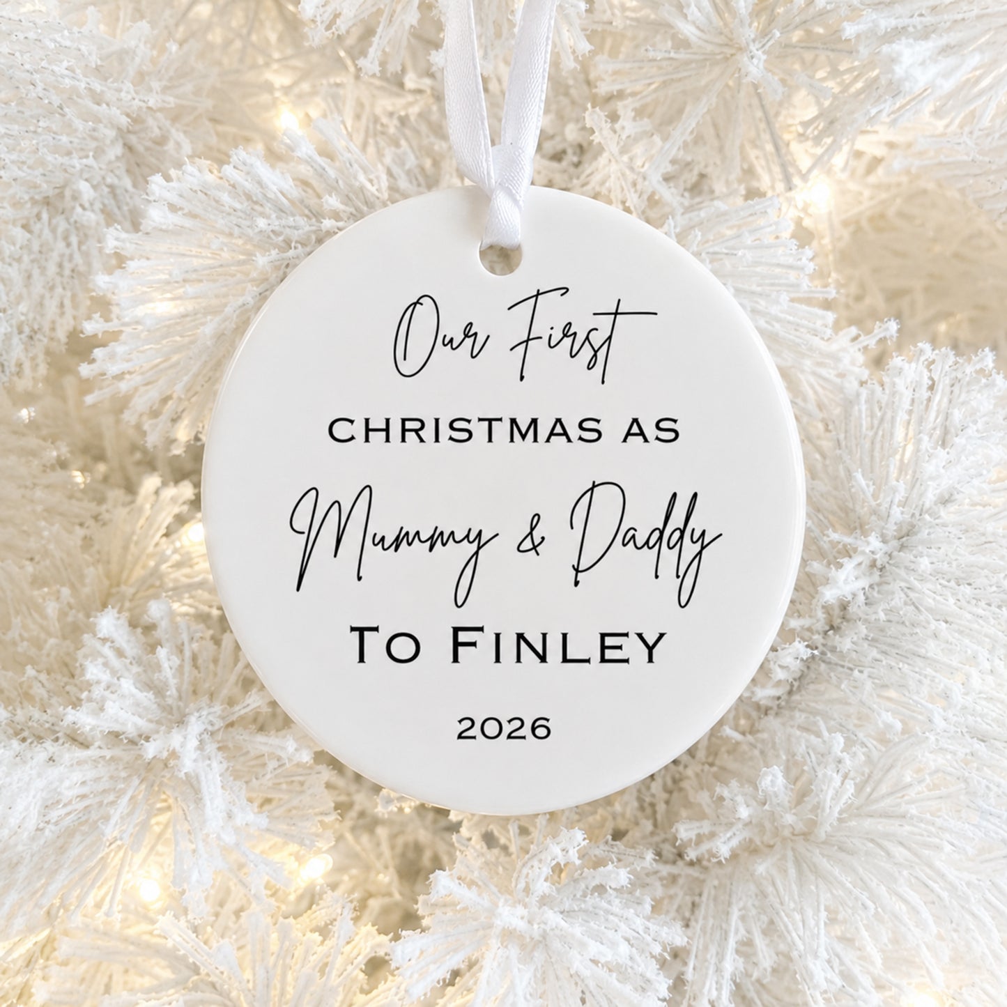 A round white ceramic Christmas ornament hanging on a frosted white Christmas tree with warm string lights. The ornament is printed with the personalized text 'Our First CHRISTMAS AS Mummy & Daddy TO FINLEY 2026' in black lettering.