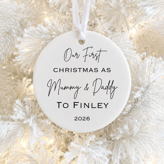 A round white ceramic Christmas ornament hanging on a frosted white Christmas tree with warm string lights. The ornament is printed with the personalized text 'Our First CHRISTMAS AS Mummy & Daddy TO FINLEY 2026' in black lettering.