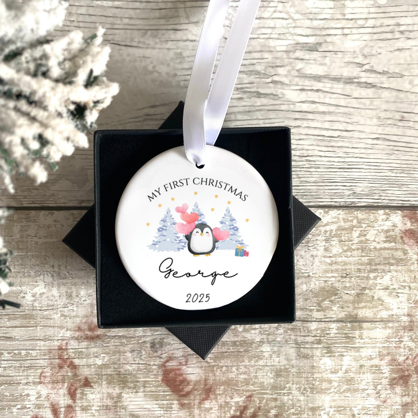 Personalized Christmas ornament with penguin design and text, placed in a black box on a wooden surface.