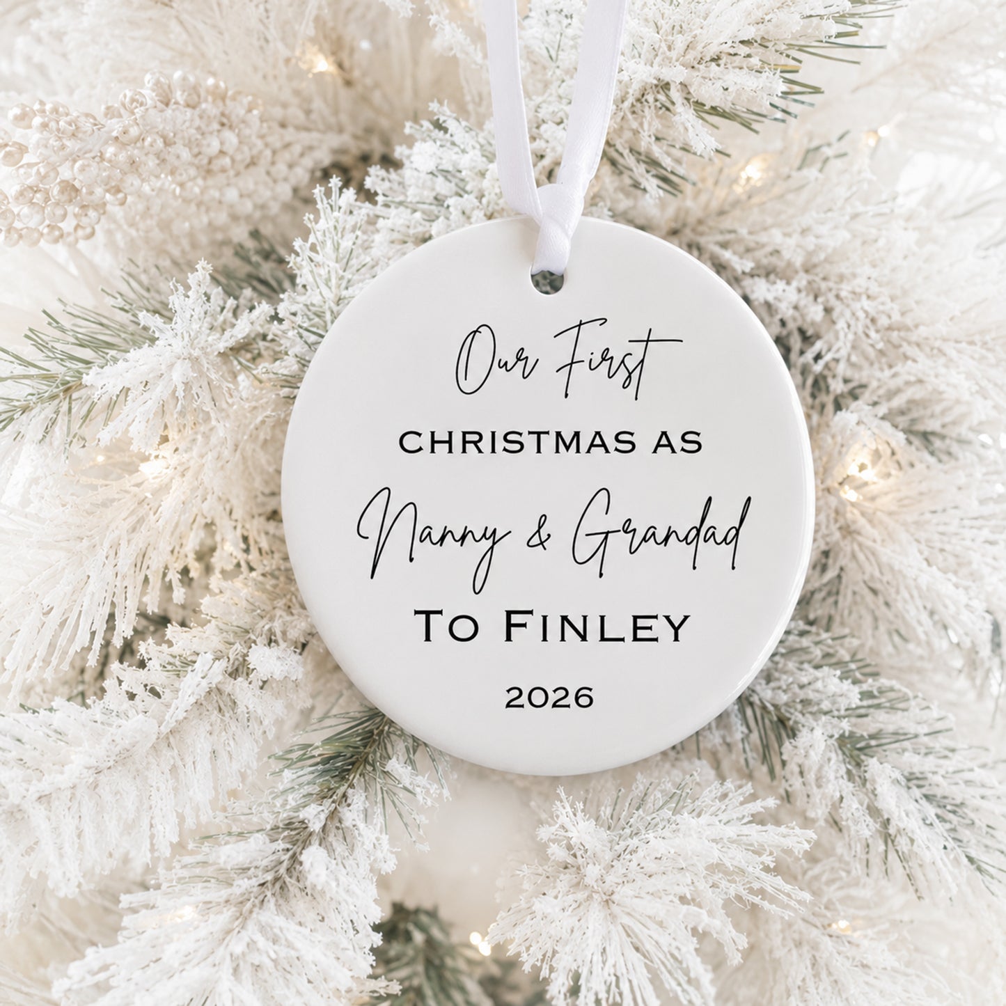 A round white personalized Christmas ornament hanging on a frosted white Christmas tree, printed with the text 'Our First CHRISTMAS AS Nanny & Grandad To Finley 2026' in black script and serif fonts.