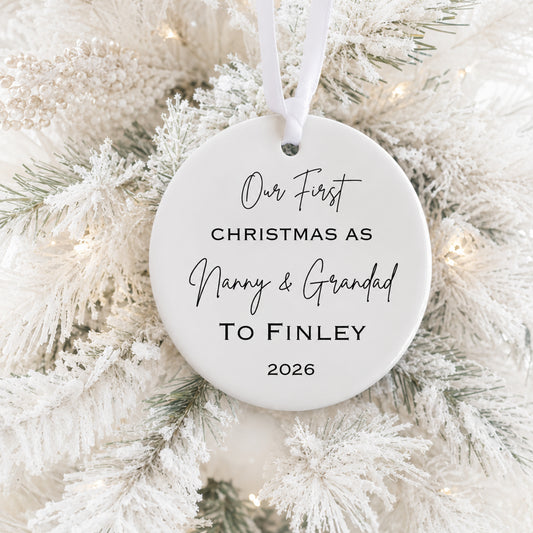 A round white personalized Christmas ornament hanging on a frosted white Christmas tree, printed with the text 'Our First CHRISTMAS AS Nanny & Grandad To Finley 2026' in black script and serif fonts.
