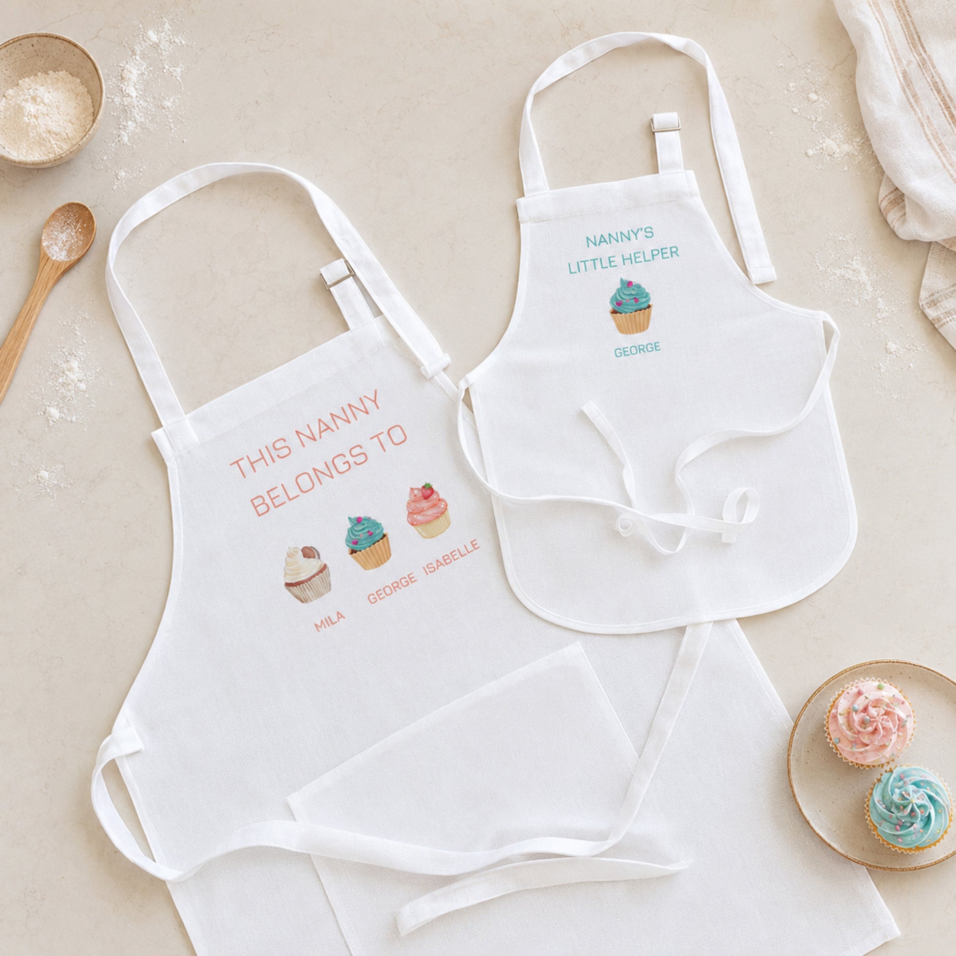 Two white aprons laid flat on a light beige surface with baking-themed props. One apron reads 'THIS NANNY BELONGS TO' with cupcake illustrations and names below, and the other reads 'NANNY'S LITTLE HELPER' with a small cupcake graphic and the name GENNAINE.