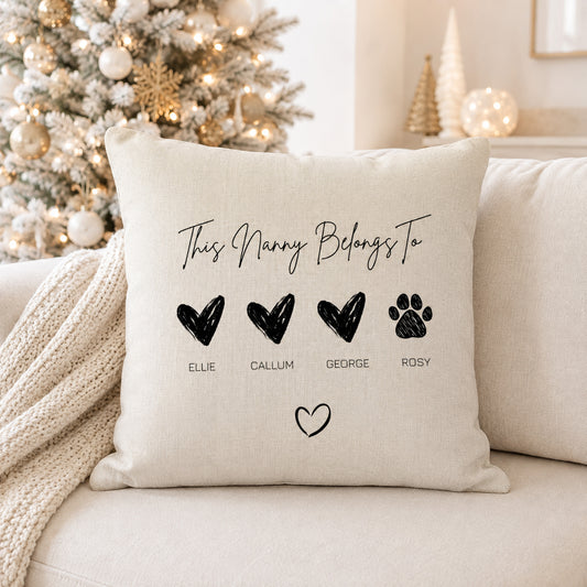 A square beige throw pillow with black printed text reading 'This Nanny Belongs To' and four black heart/paw icons labeled Ellie, Callum, George, and Rosy, styled on a cream sofa with a knitted blanket and a decorated Christmas tree in the background.