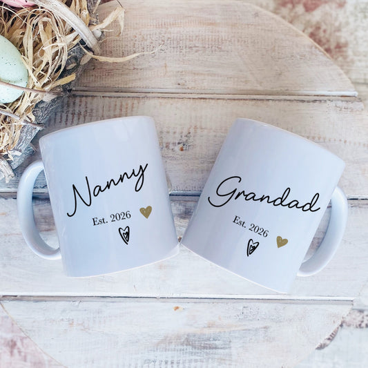 Two mugs labeled 'Nanny' and 'Grandad' with decorative eggs and nest on a wooden surface.