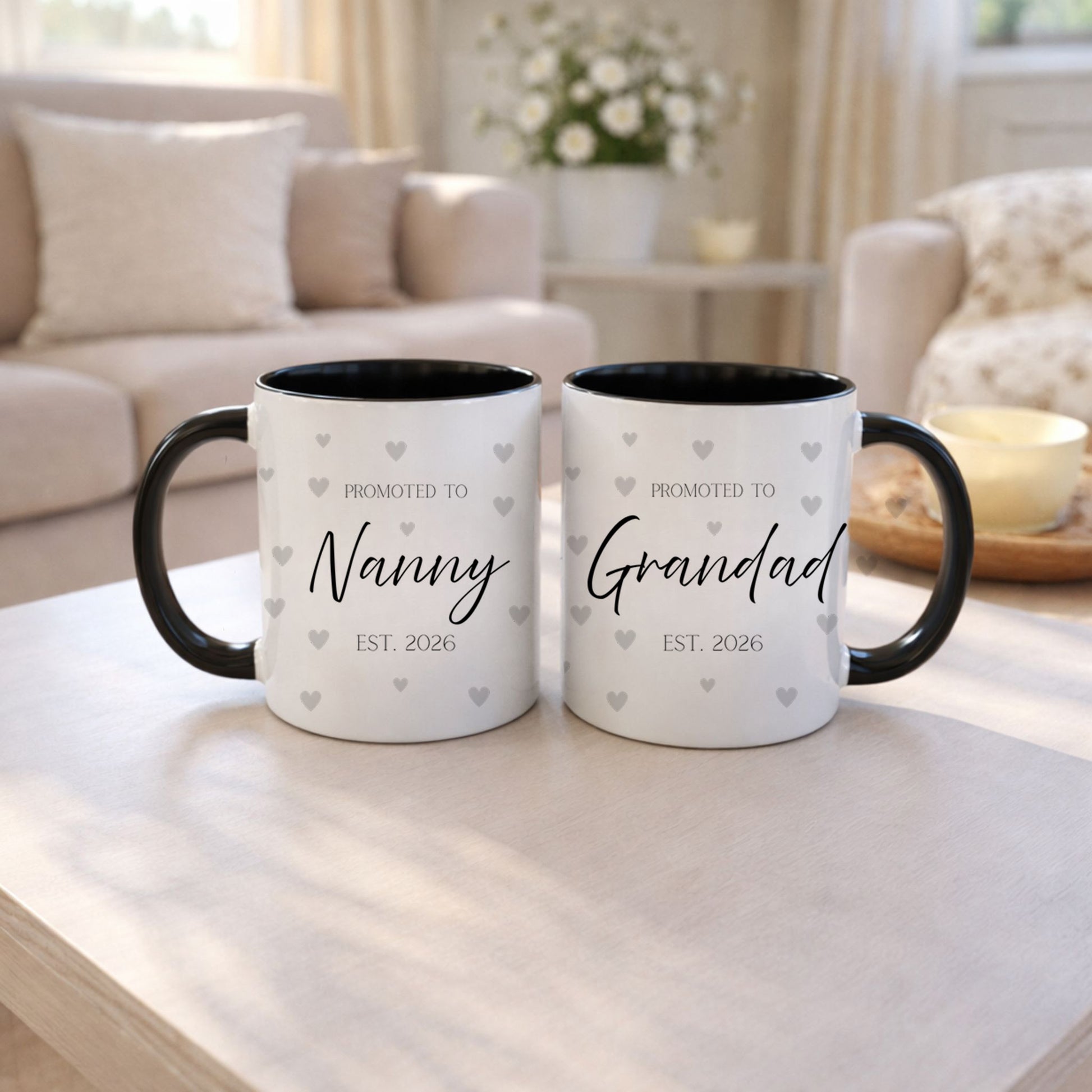 Two mugs with 'Promoted to Nanny EST. 2026' and 'Promoted to Grandad EST. 2026' text on a table in a living room.