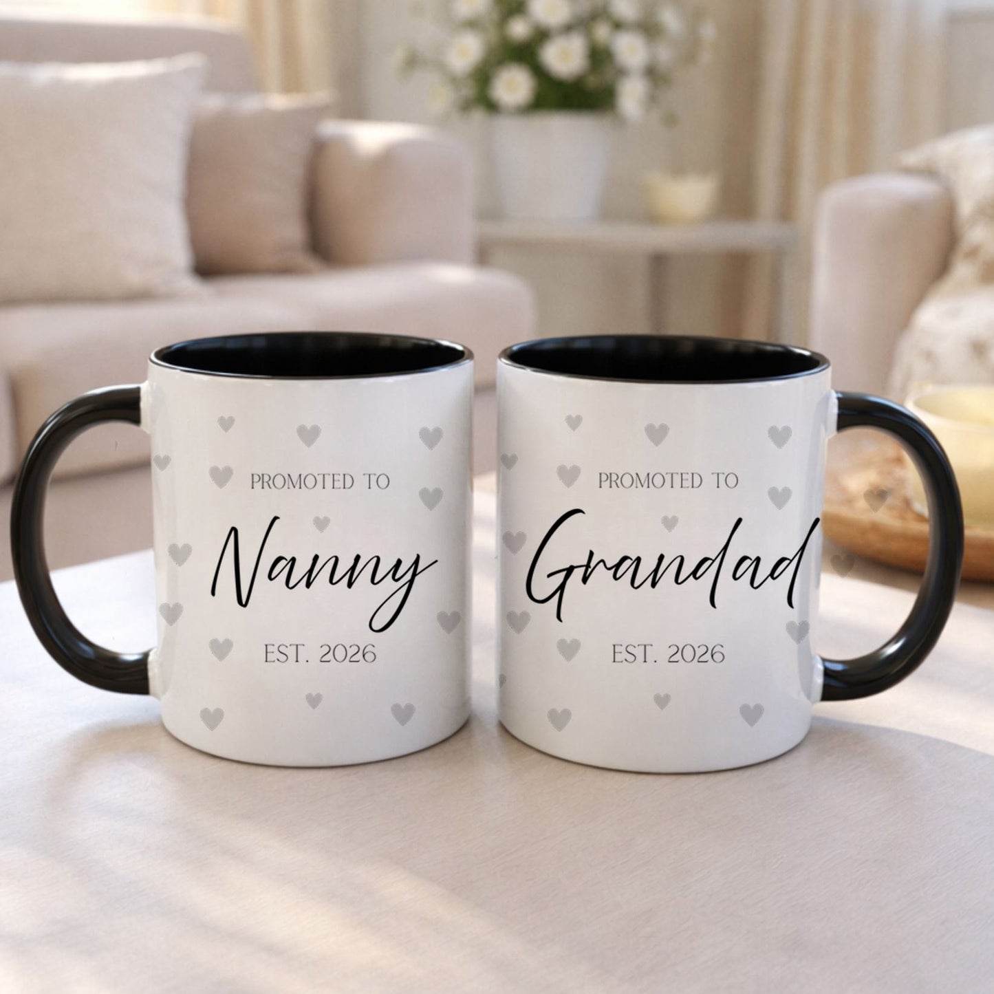 Two mugs with 'Promoted to Nanny EST. 2026' and 'Promoted to Grandad EST. 2026' text on a table in a living room.