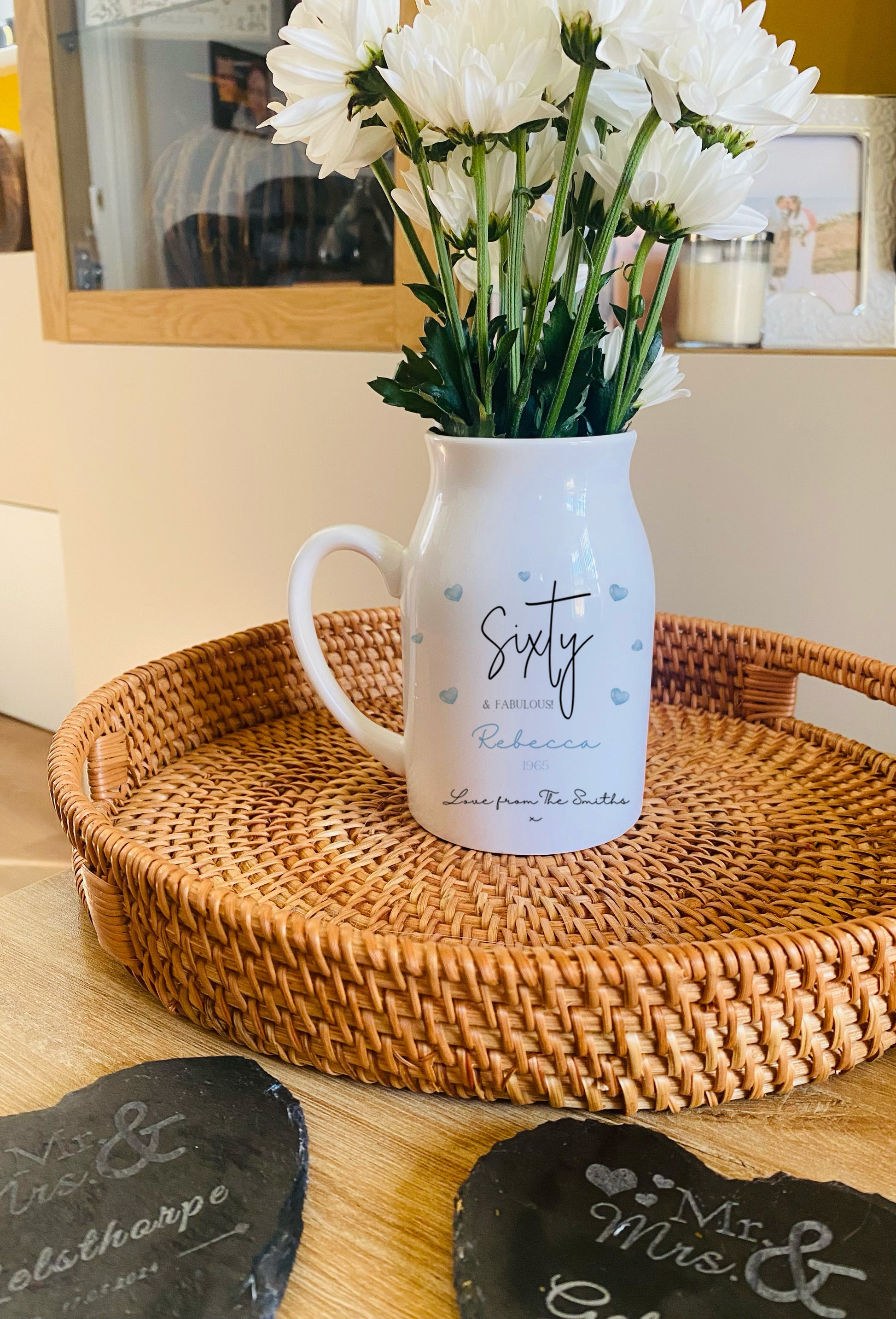 White pitcher vase with 'Sixty' text on a woven tray with flowers