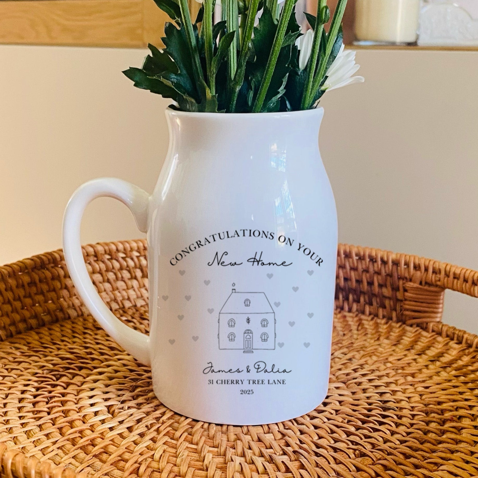 A personalised White pitcher vase with flowers on a woven tray in a home setting. The wording includes 'Congratulations on your new home'
