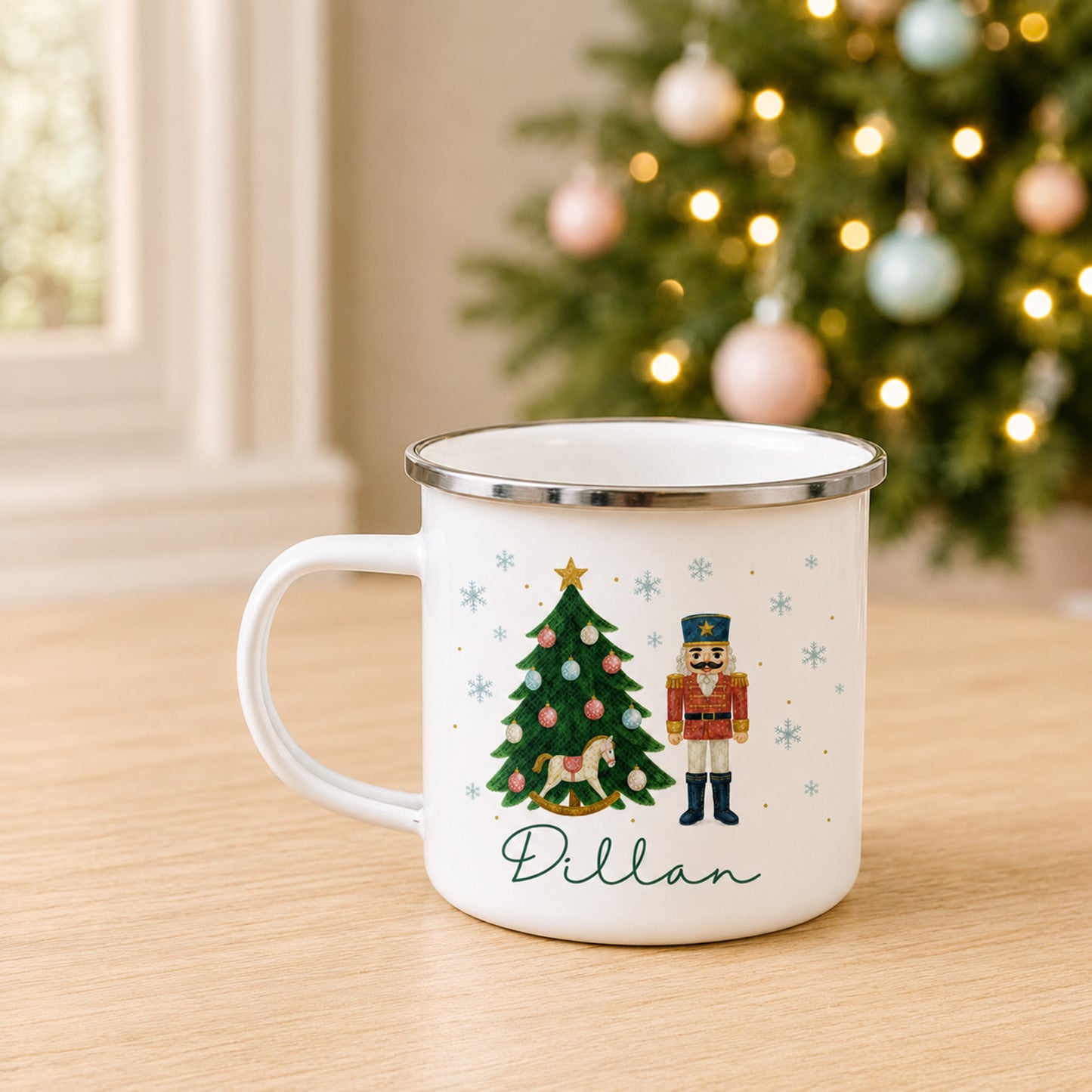 A white enamel-style mug with a silver rim sits on a wooden table in a cozy indoor setting, with a blurred Christmas tree in the background. The mug features a printed Christmas tree, a nutcracker, snowflakes, and the name 'Dillan' in green script.