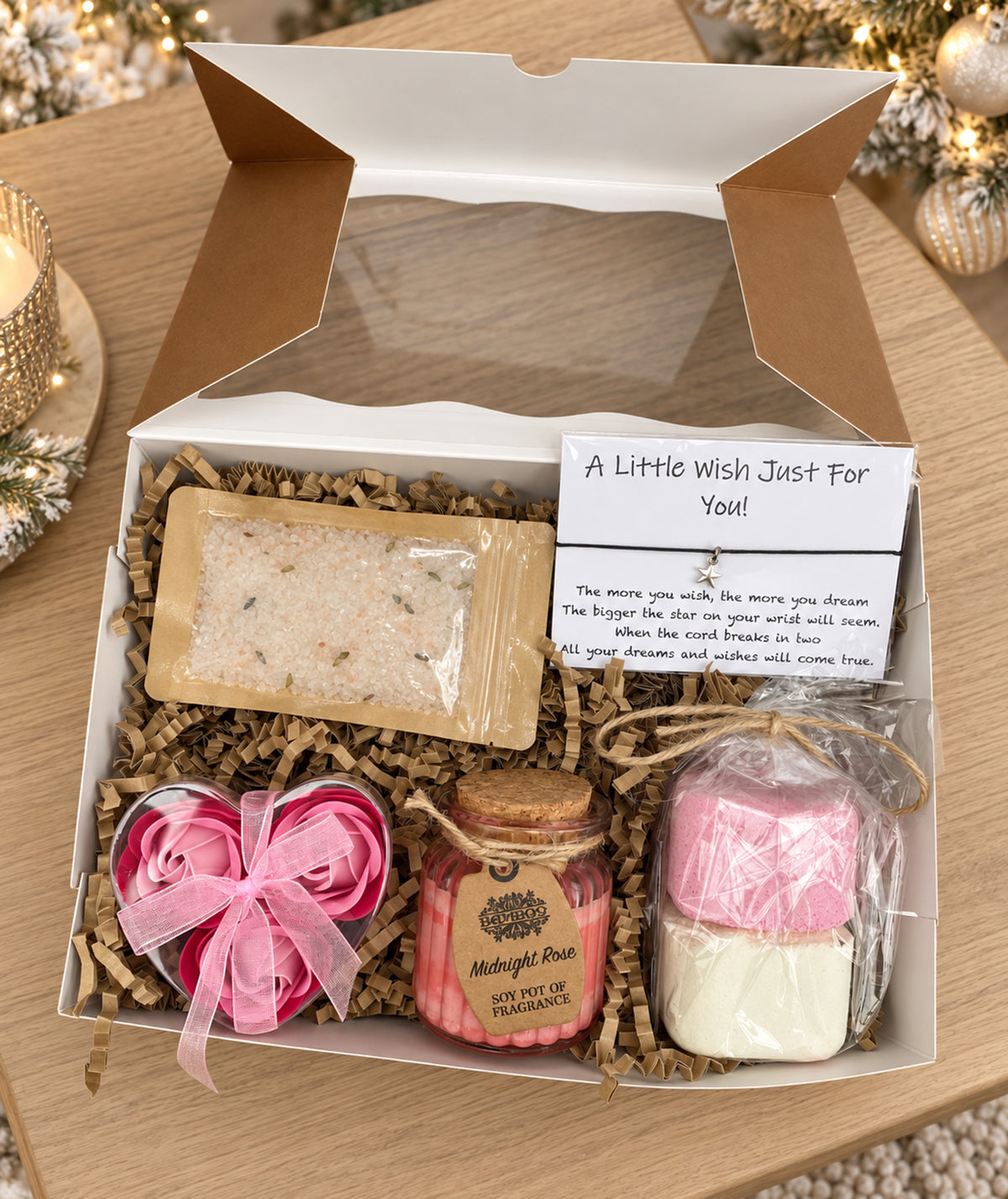 A Christmas-themed gift set in a brown kraft box with a lace ribbon, containing shower steamers, a pink heart-shaped soap flowers, a small candle and bath salts, displayed on a wooden table with warm holiday lights in the background.