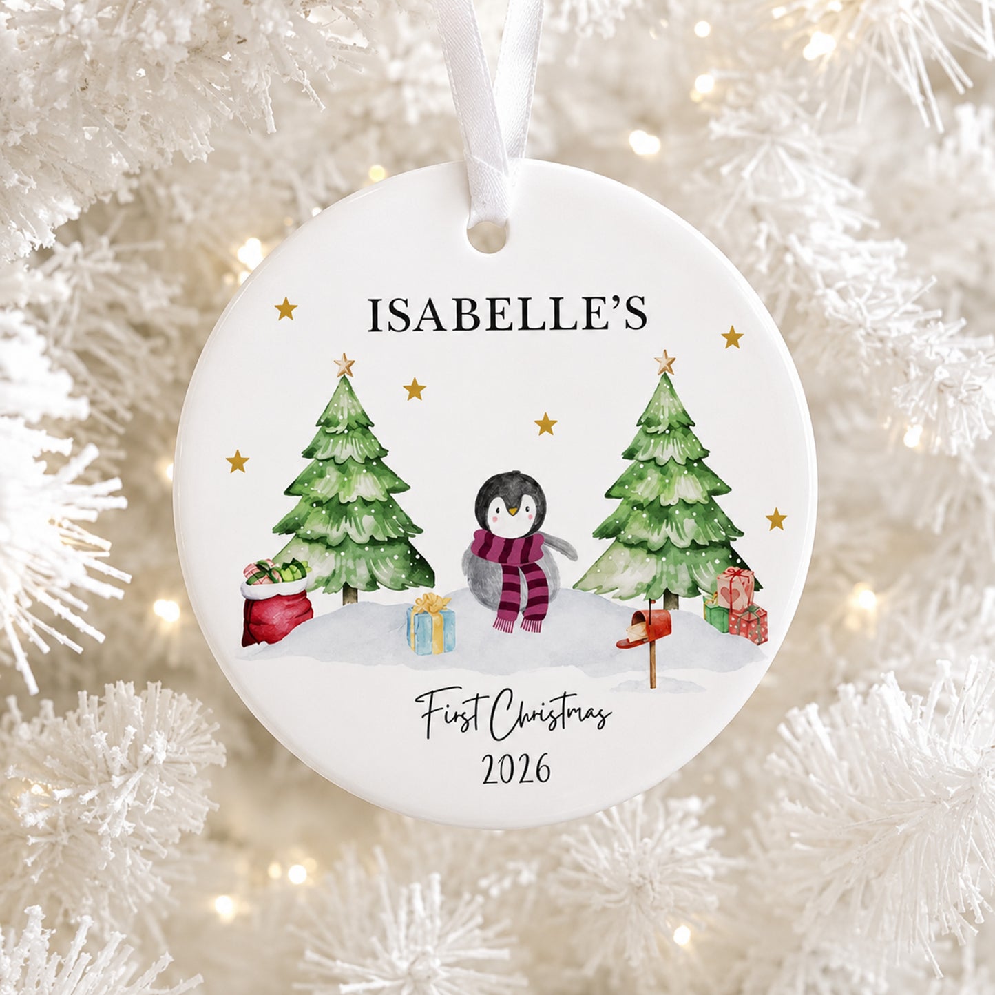 A round white personalized Christmas ornament hanging on a white tree with warm string lights. The ornament features a printed illustration of a penguin in a red and purple striped scarf between two green Christmas trees, with the text 'ISABELLE'S First Christmas 2026'.