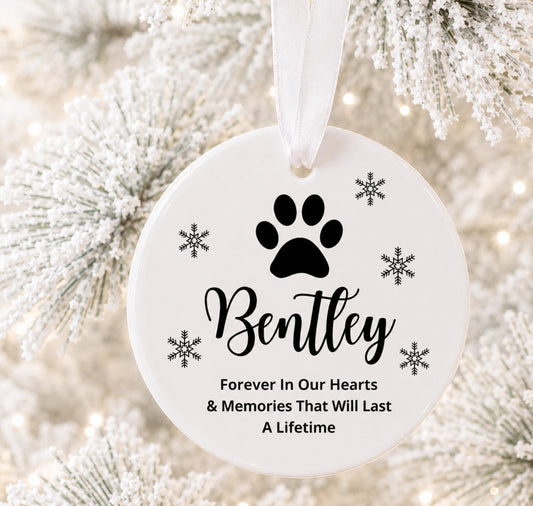 A round white pet memorial Christmas ornament hanging from a white ribbon, printed with a black paw print, the name 'Bentley,' snowflakes, and the text 'Forever In Our Hearts & Memories That Will Last A Lifetime,' shown against frosted tree branches with soft lights.