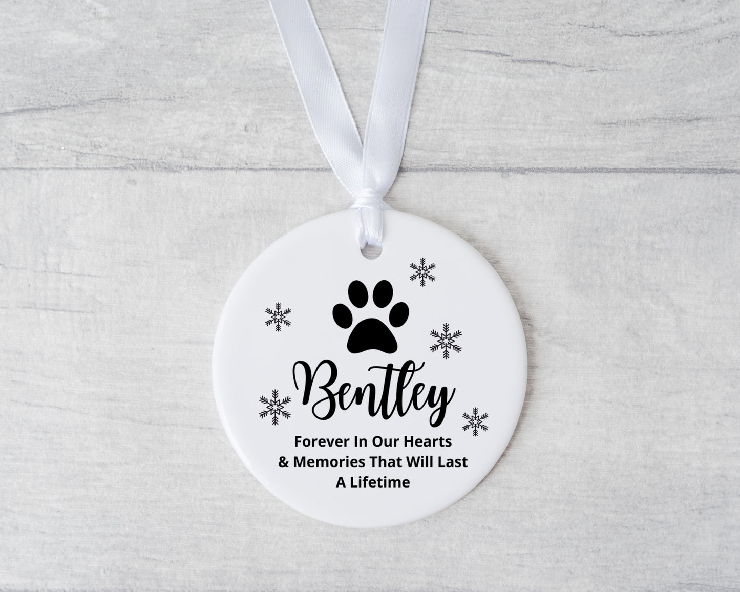 Round ceramic ornament with paw print and text on a white ribbon against a light gray background