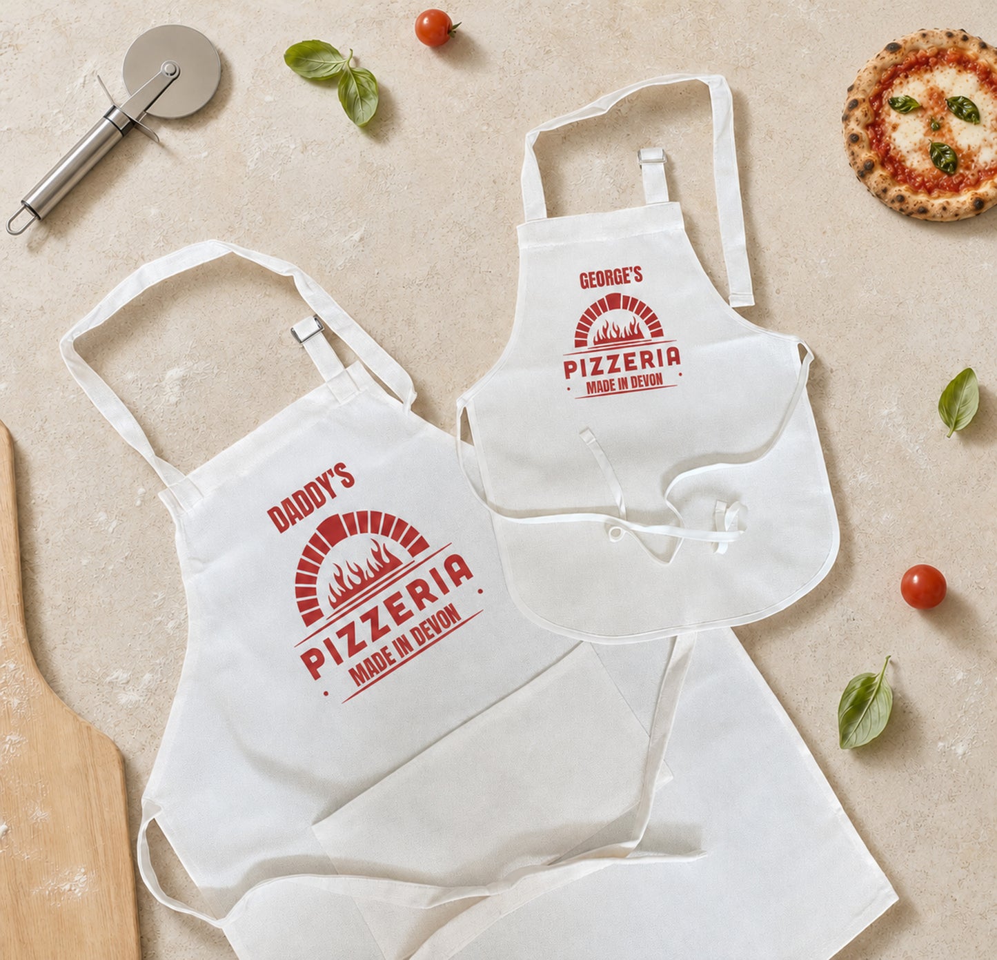Two white fabric aprons laid flat on a light beige surface, each printed with a red pizza-themed logo and text. One apron reads "Daddy's Pizzeria Made in Love" and the other reads "George's Pizzeria Made in Love.