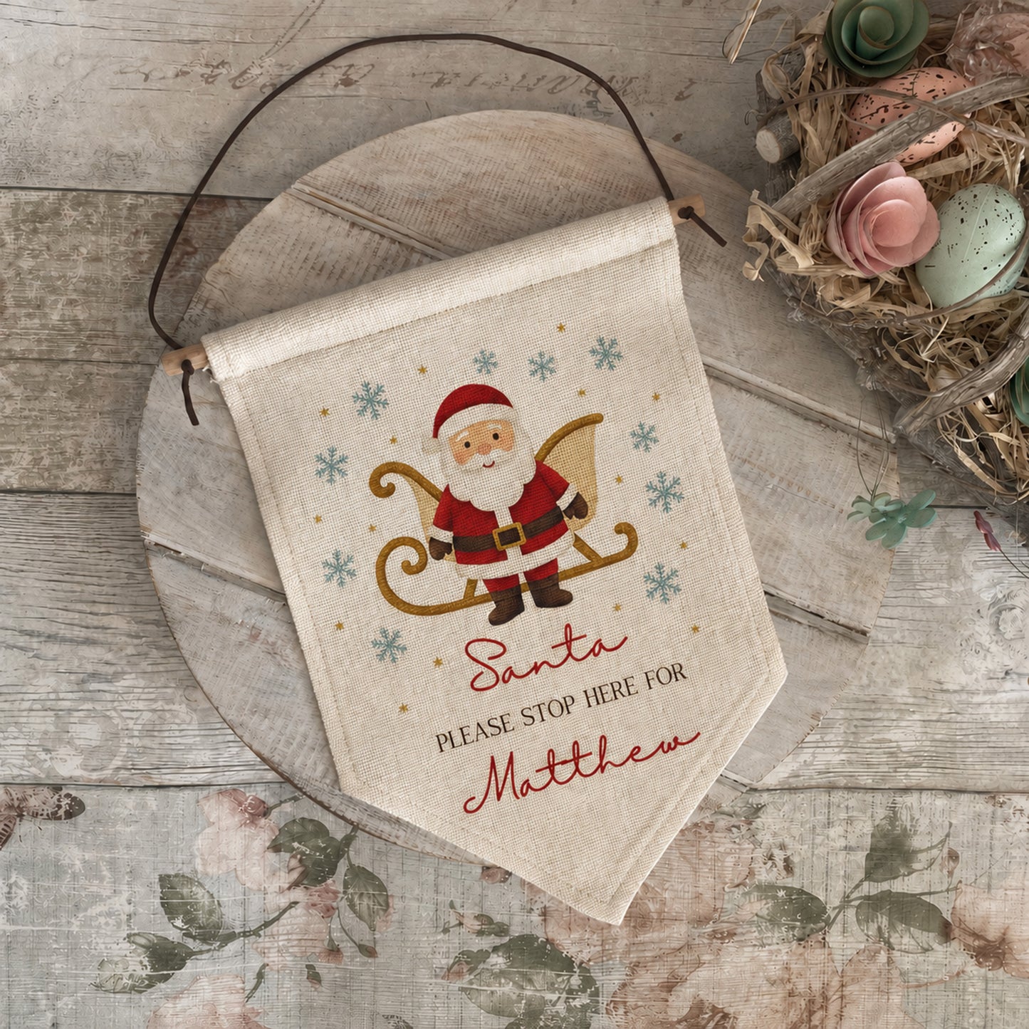 A personalized canvas hanging banner with a Santa Claus design, laid on a rustic wooden surface with a decorative bird nest nearby.