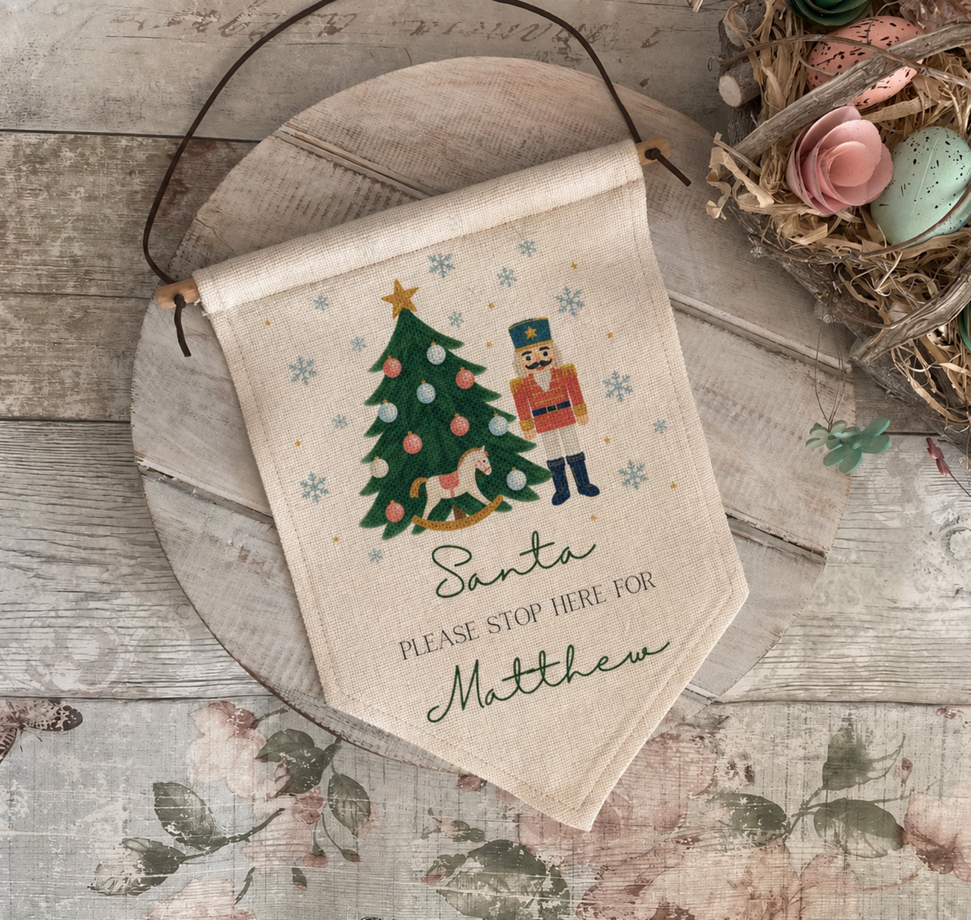 A personalized canvas hanging banner with a Christmas nutcracker and tree design, laid on a rustic wooden surface with a decorative bird nest nearby.