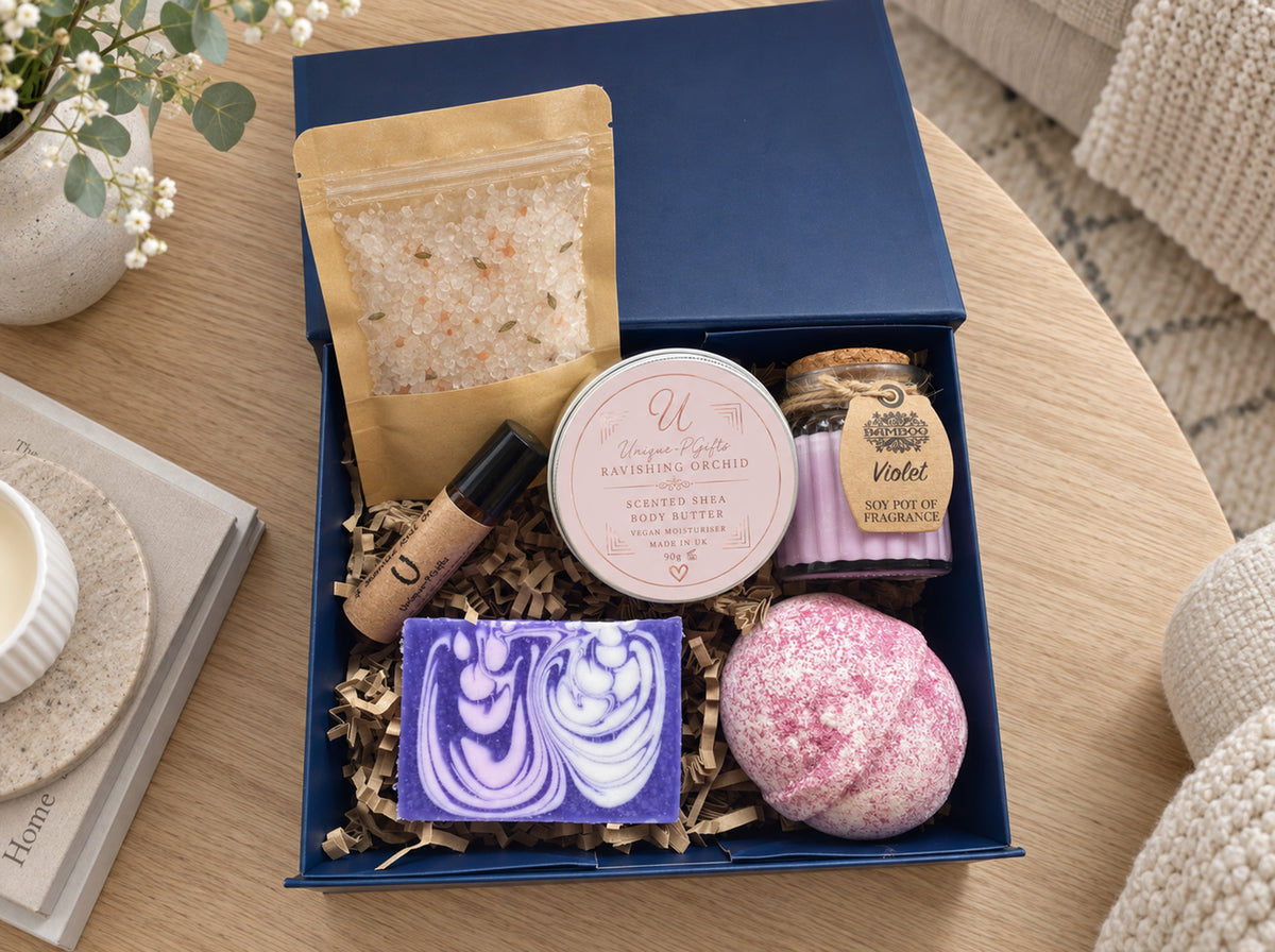 A curated bath and body gift box arranged in a navy blue tray on a wooden table. The set includes a lavender and white marbled soap bar, a pink bath bomb, a round tin labeled 'Scented Shea Hand Butter', a small jar labeled 'Violet', a gold pouch of bath salts, and a black-and-white stick bottle, all nestled in shredded paper filler.