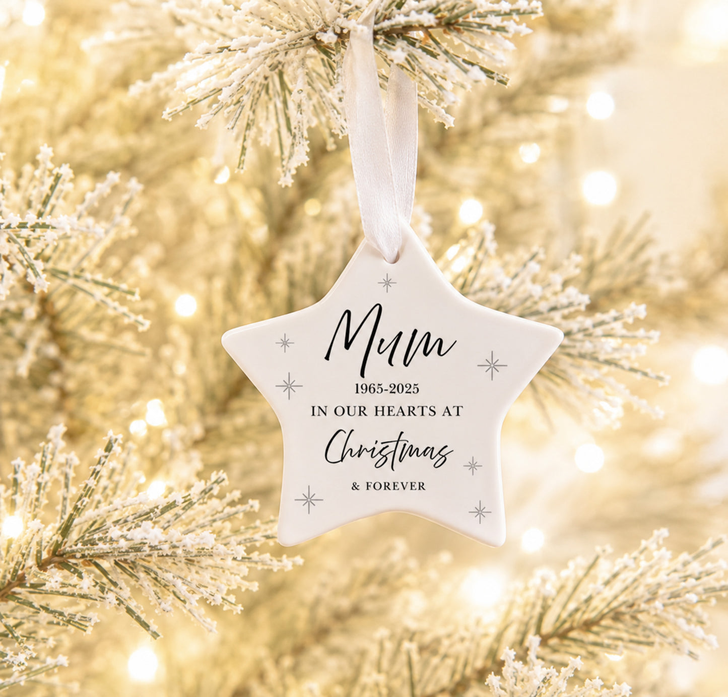 A white star-shaped hanging ornament with printed memorial text and small silver snowflake motifs, suspended from a white ribbon on a frosted Christmas tree branch with warm bokeh lights in the background.