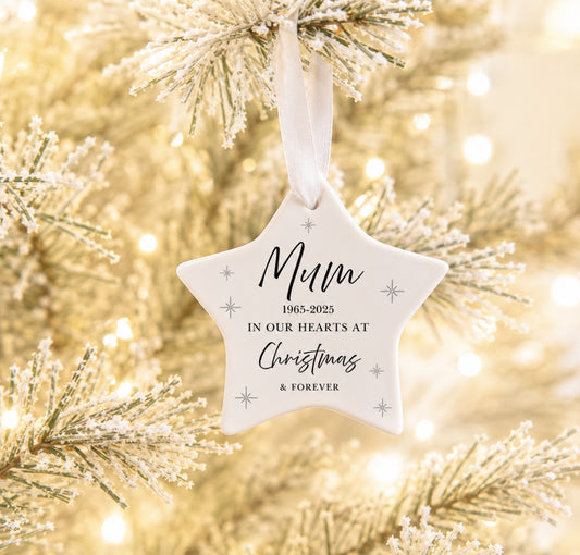 A white star-shaped hanging ornament with printed memorial text and small silver snowflake motifs, suspended from a white ribbon on a frosted Christmas tree branch with warm bokeh lights in the background.