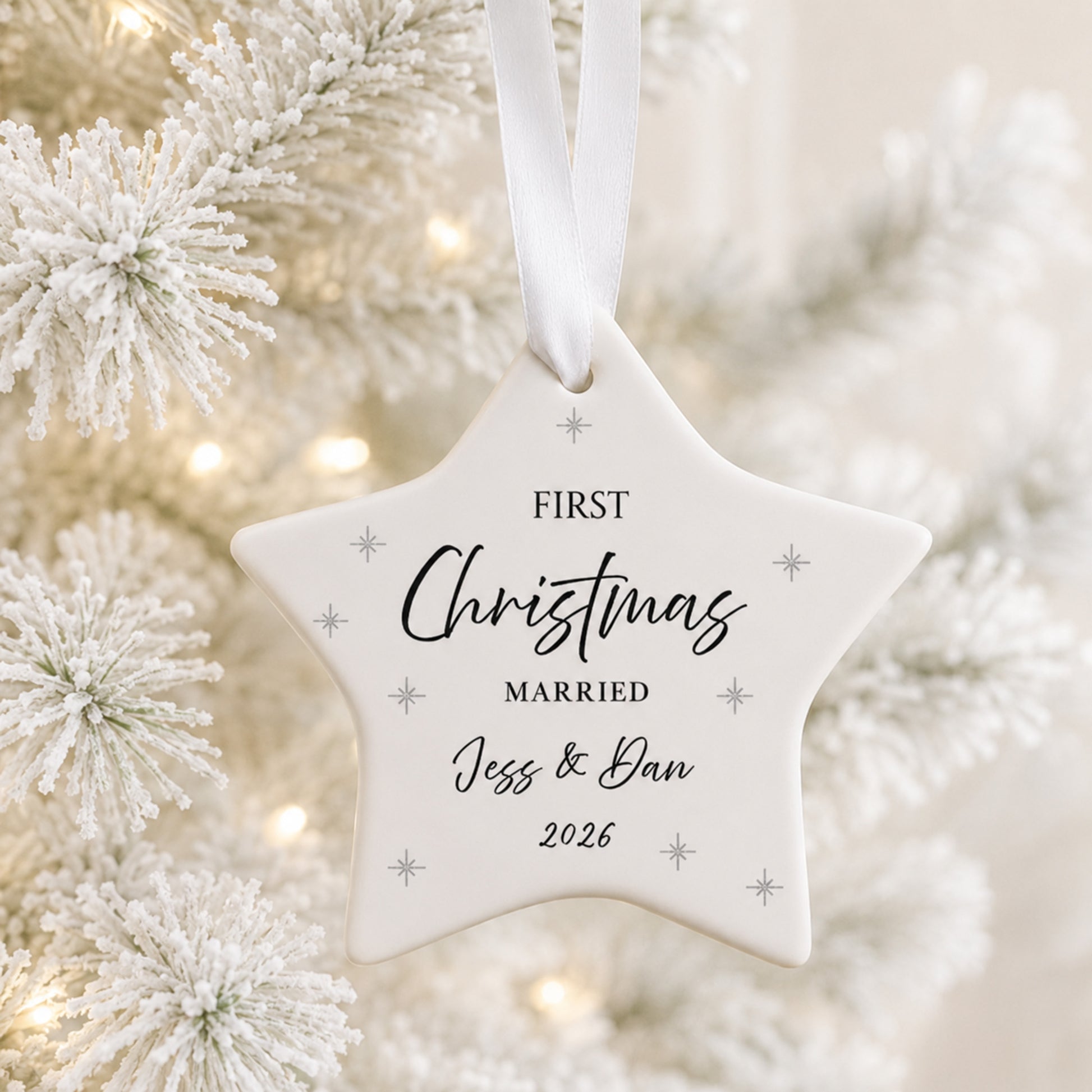 A white star-shaped Christmas ornament hanging from a white ribbon, printed with the words 'FIRST Christmas MARRIED Jess & Dan 2026' in black script and serif fonts, with small silver star accents. The ornament is photographed against a softly blurred festive background of white frosted pine branches and warm bokeh lights.