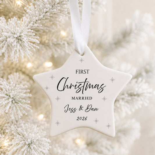 A white star-shaped Christmas ornament hanging from a white ribbon, printed with the words 'FIRST Christmas MARRIED Jess & Dan 2026' in black script and serif fonts, with small silver star accents. The ornament is photographed against a softly blurred festive background of white frosted pine branches and warm bokeh lights.
