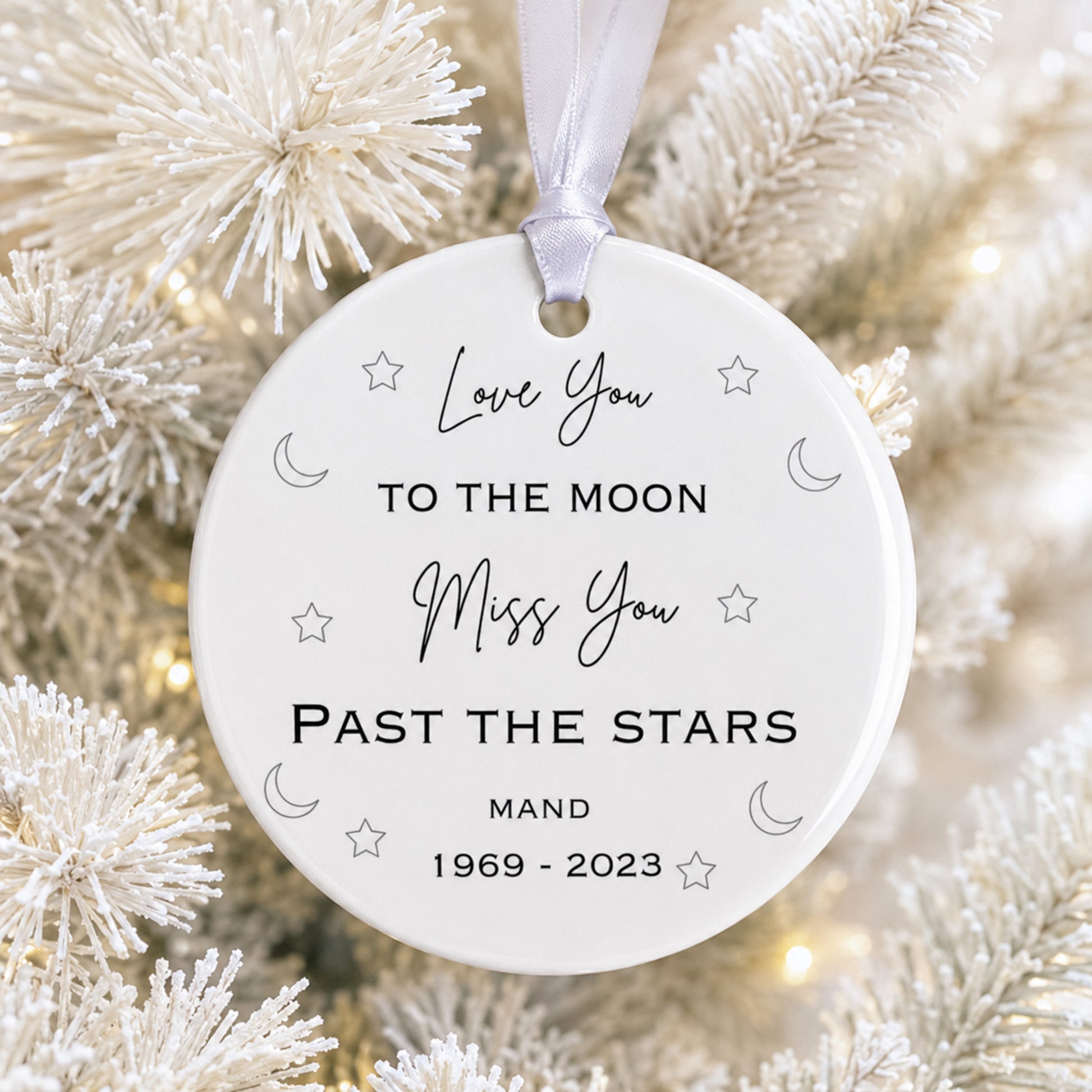 A round white memorial Christmas ornament hanging from a white ribbon, printed with the words 'Love You TO THE MOON Miss You PAST THE STARS' and 'MAND 1969 - 2023', surrounded by small moon, star, and heart motifs. The ornament is photographed among white frosted tree branches with warm blurred lights in the background.