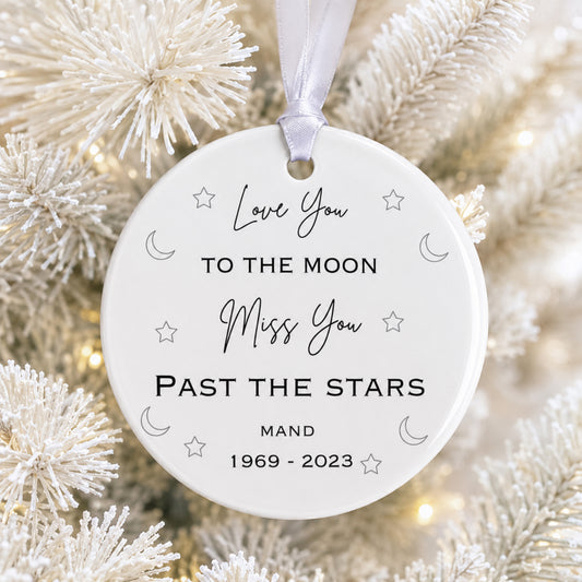 A round white memorial Christmas ornament hanging from a white ribbon, printed with the words 'Love You TO THE MOON Miss You PAST THE STARS' and 'MAND 1969 - 2023', surrounded by small moon, star, and heart motifs. The ornament is photographed among white frosted tree branches with warm blurred lights in the background.
