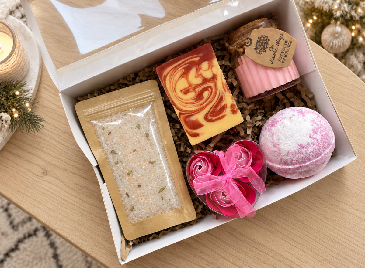 A curated gift box containing a marbled soap bar, a pink bath bomb, a pink rose-shaped item tied with a ribbon, a pink bath bomb, and a clear pouch of bath salts, all arranged in shredded paper inside an open white box on a wooden surface.