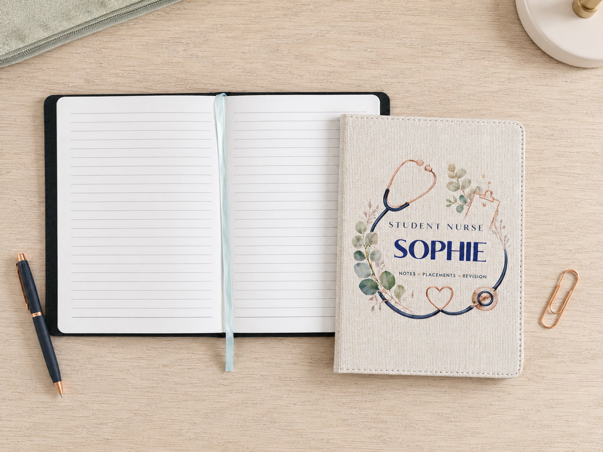 A personalized student nurse notebook cover with a stethoscope and floral design reading 'Student Nurse Sophie' is shown on a light wood desk next to an open lined notebook, a pen, and a paperclip.