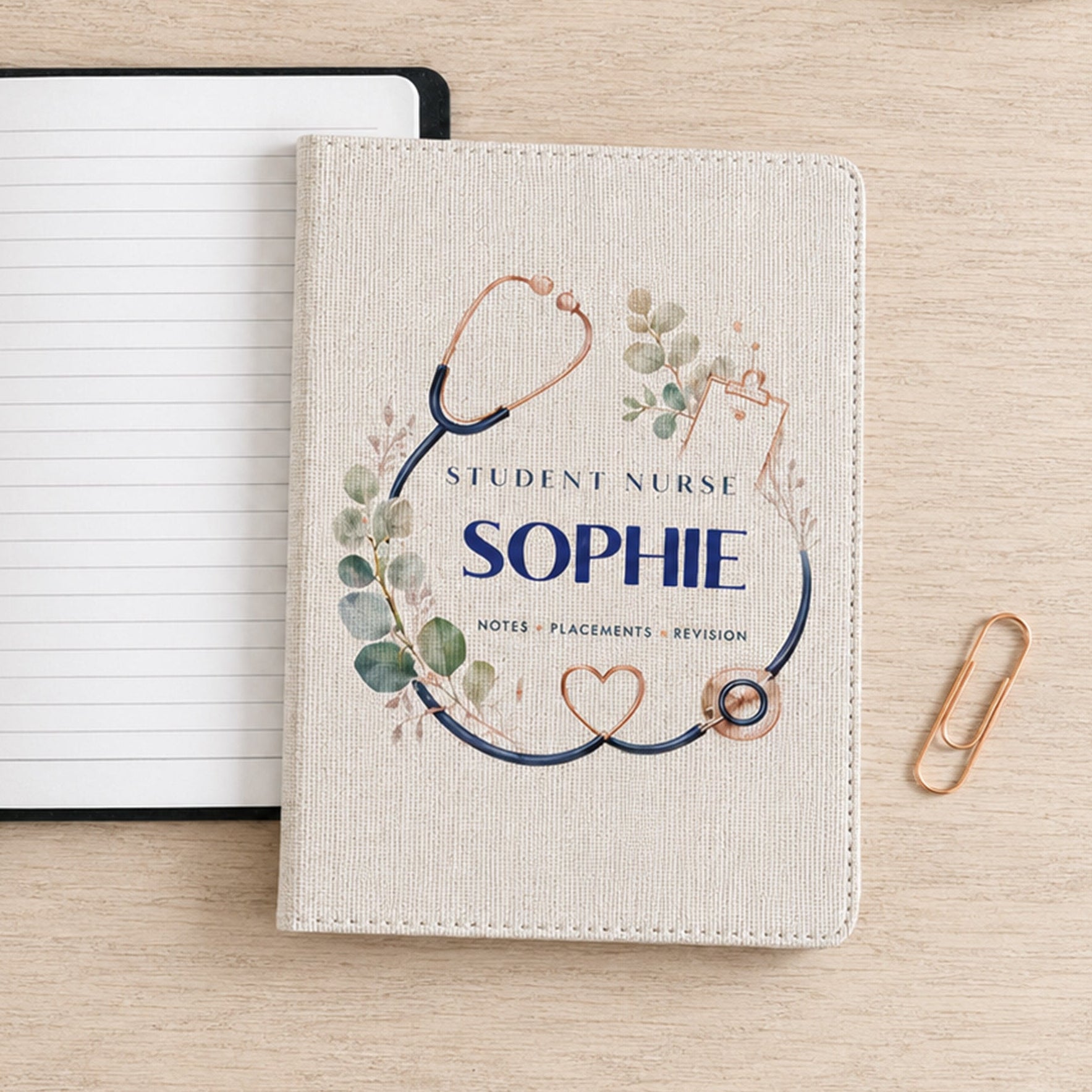 A personalized student nurse notebook cover with a stethoscope and floral design reading 'Student Nurse Sophie' is shown on a light wood desk next to an open lined notebook, a pen, and a paperclip.