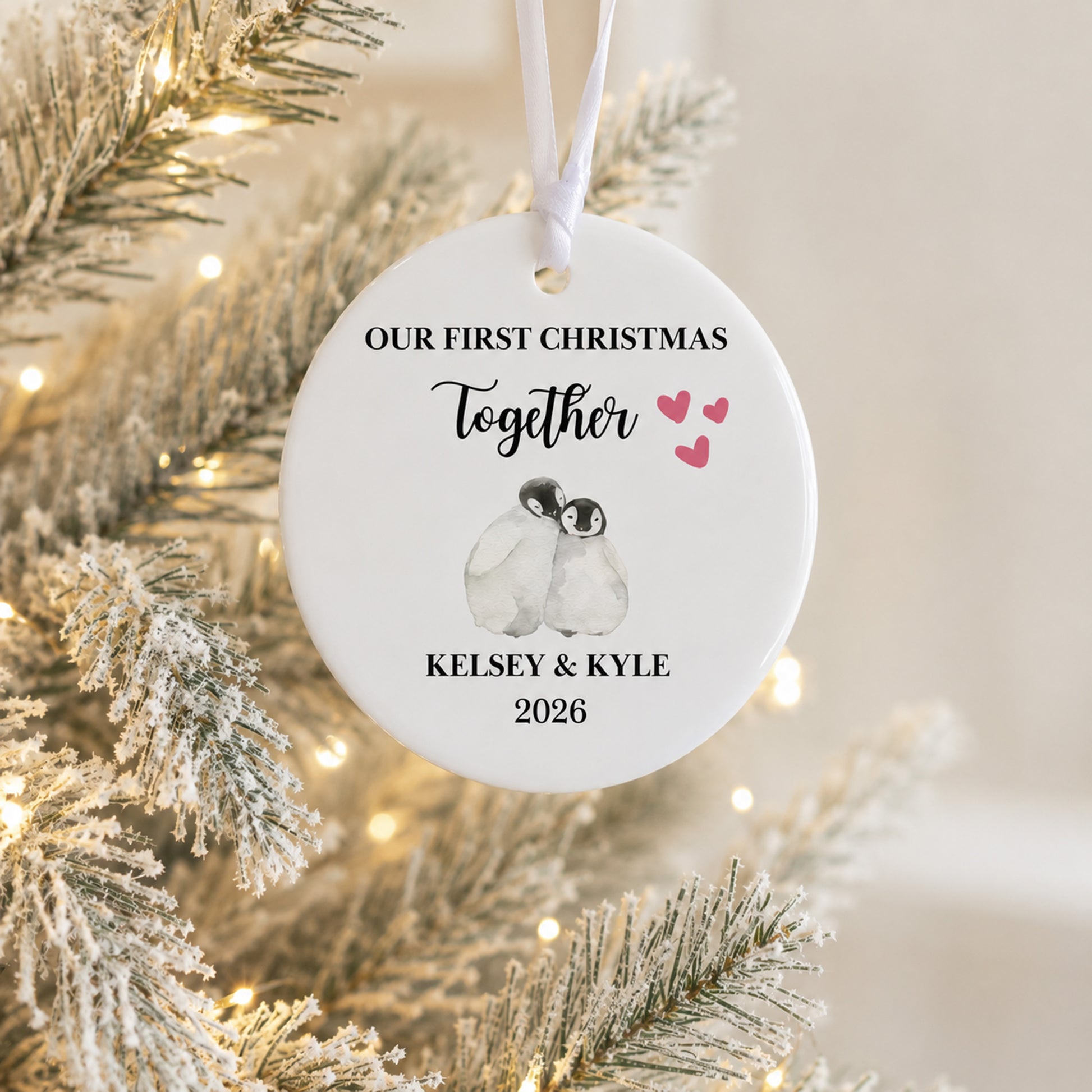 A personalized round white Christmas ornament hanging from a white ribbon, featuring a watercolor penguin illustration and the text 'OUR FIRST CHRISTMAS Together Kelsey & Kyle 2026' in black script, with small pink hearts. The ornament is photographed against a softly blurred flocked Christmas tree with warm string lights.