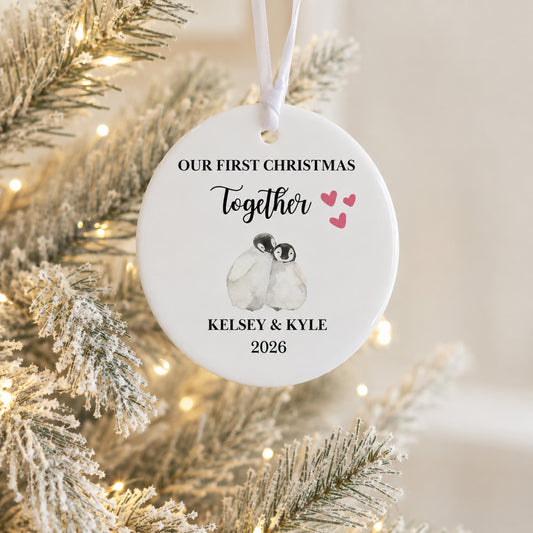 A personalized round white Christmas ornament hanging from a white ribbon, featuring a watercolor penguin illustration and the text 'OUR FIRST CHRISTMAS Together Kelsey & Kyle 2026' in black script, with small pink hearts. The ornament is photographed against a softly blurred flocked Christmas tree with warm string lights.