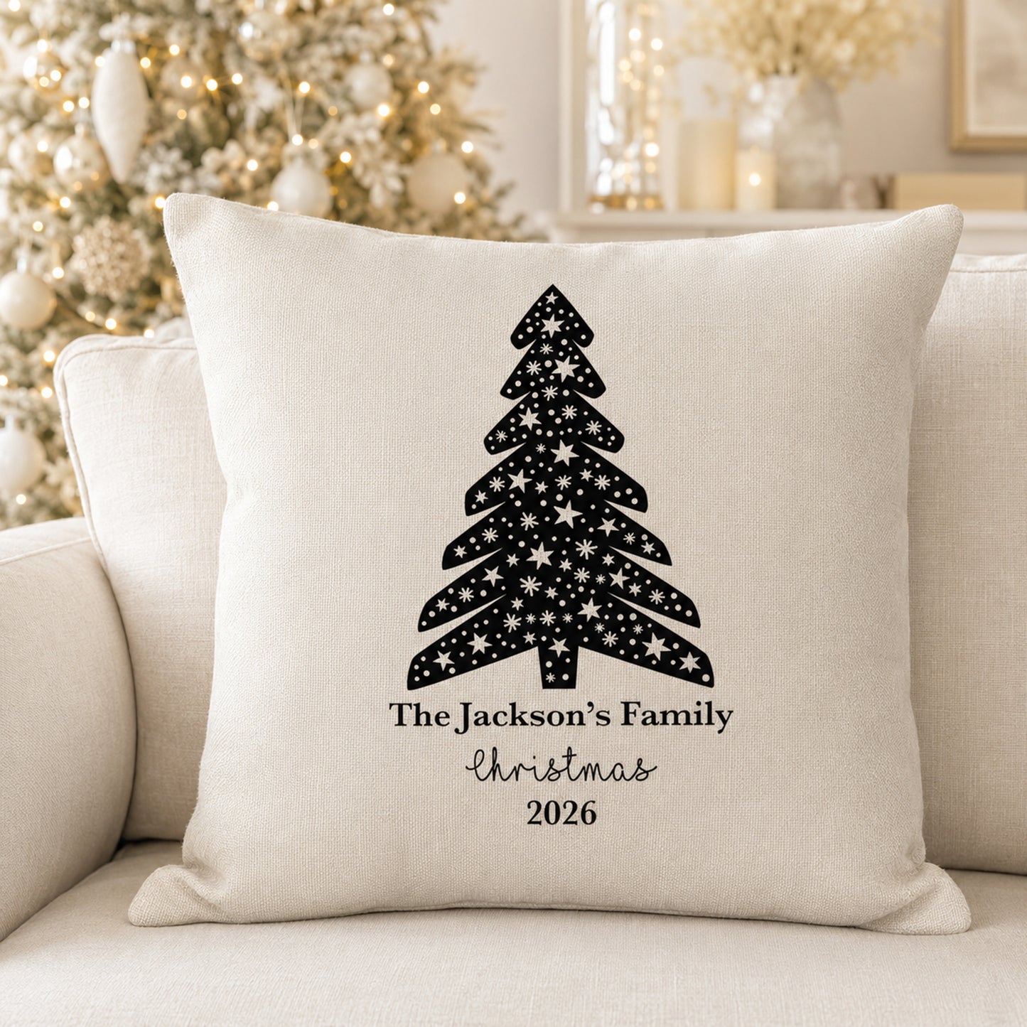 A square off-white throw pillow with a black Christmas tree design filled with white stars and the personalized text 'The Jackson's Family Christmas 2026', displayed on a beige sofa with a blurred decorated Christmas tree in the background.