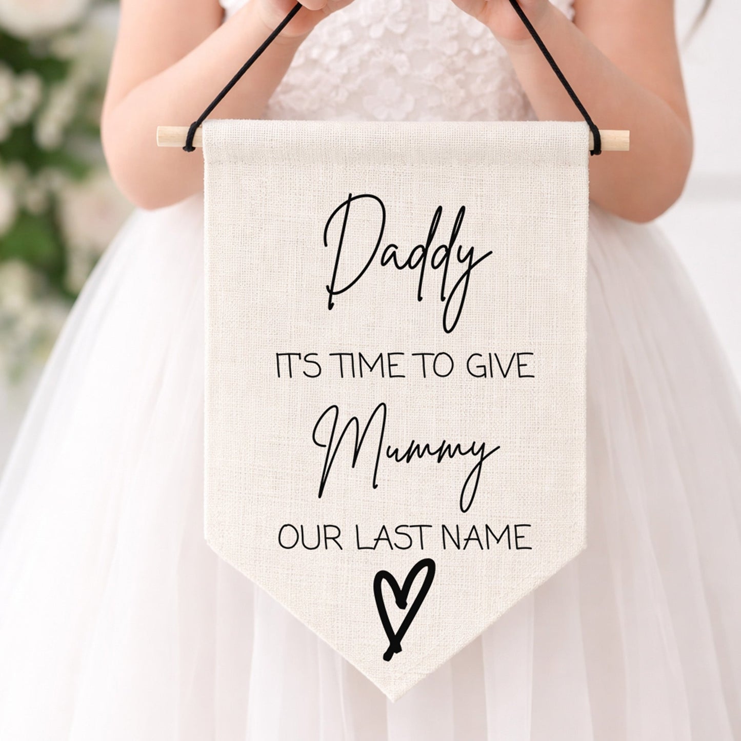 Child holding a sign with 'Daddy it's time to give mummy our last name'  in a white dress against a blurred floral background