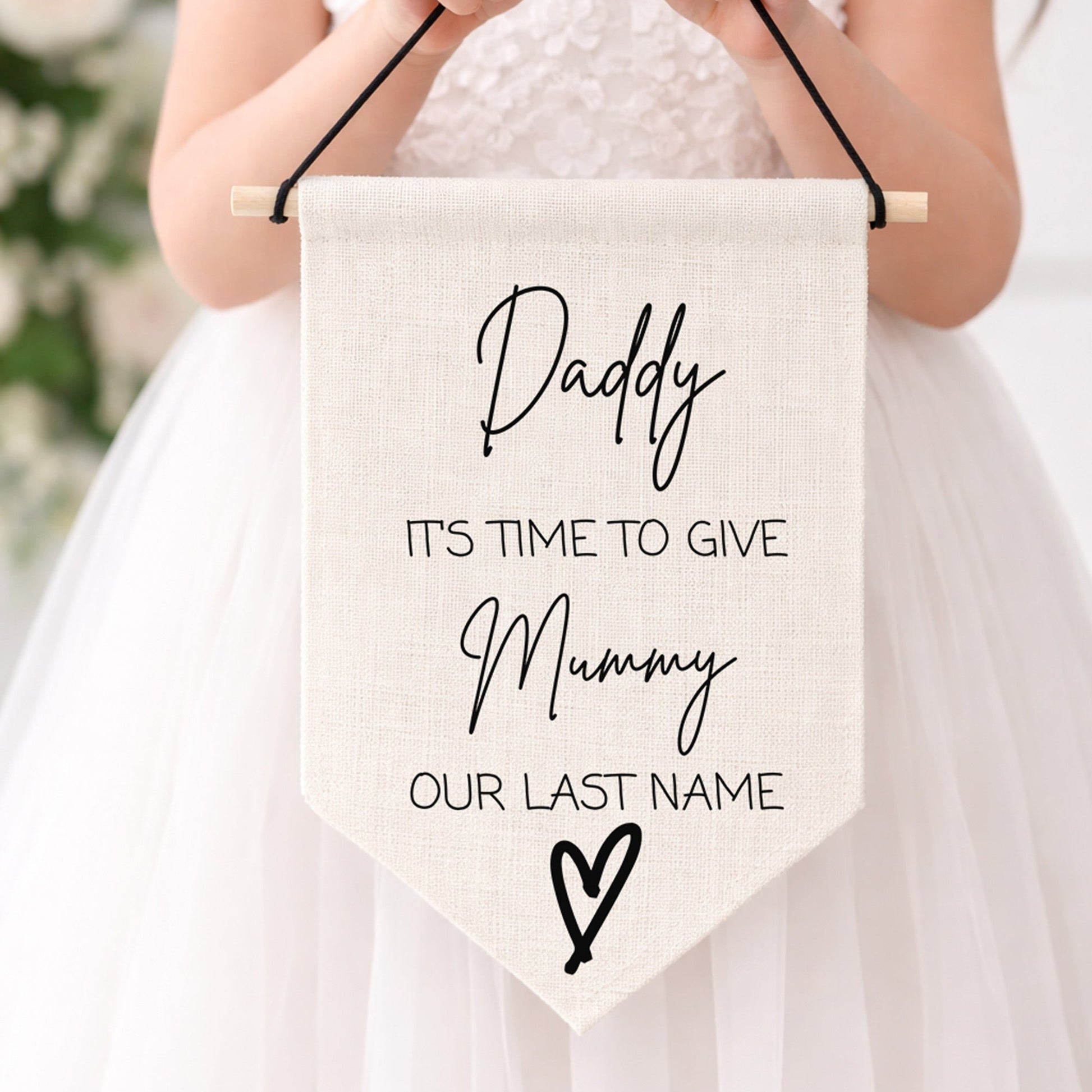 Child holding a sign with 'Daddy it's time to give mummy our last name'  in a white dress against a blurred floral background