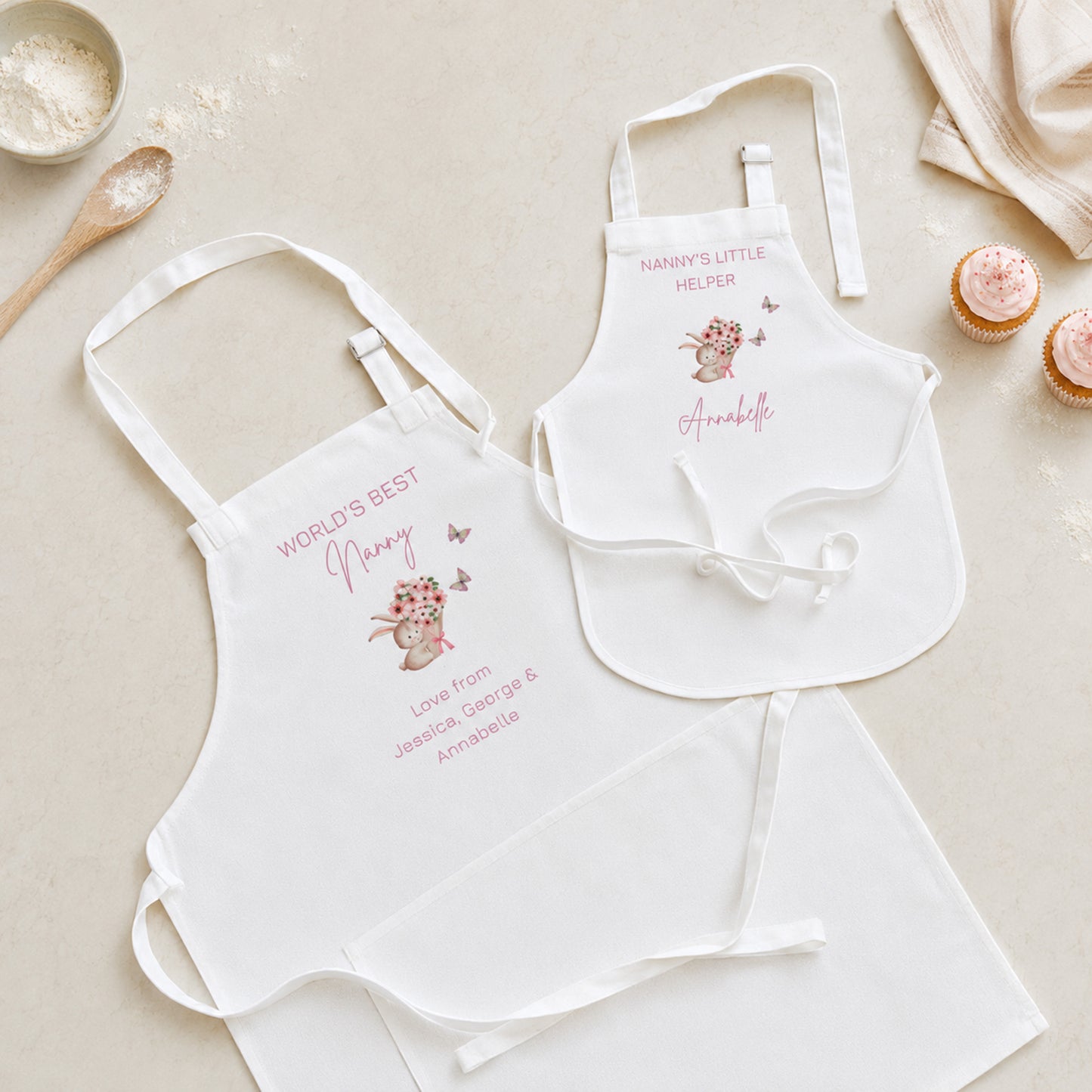 Two white baby bib aprons laid flat on a light beige surface with styled kitchen props. One bib reads "WORLD'S BEST Nanny" with a pink bunny illustration and personalized text below; the other reads "NANNY'S LITTLE MILITER" with a bunny and the name "Annabelle" in pink script.